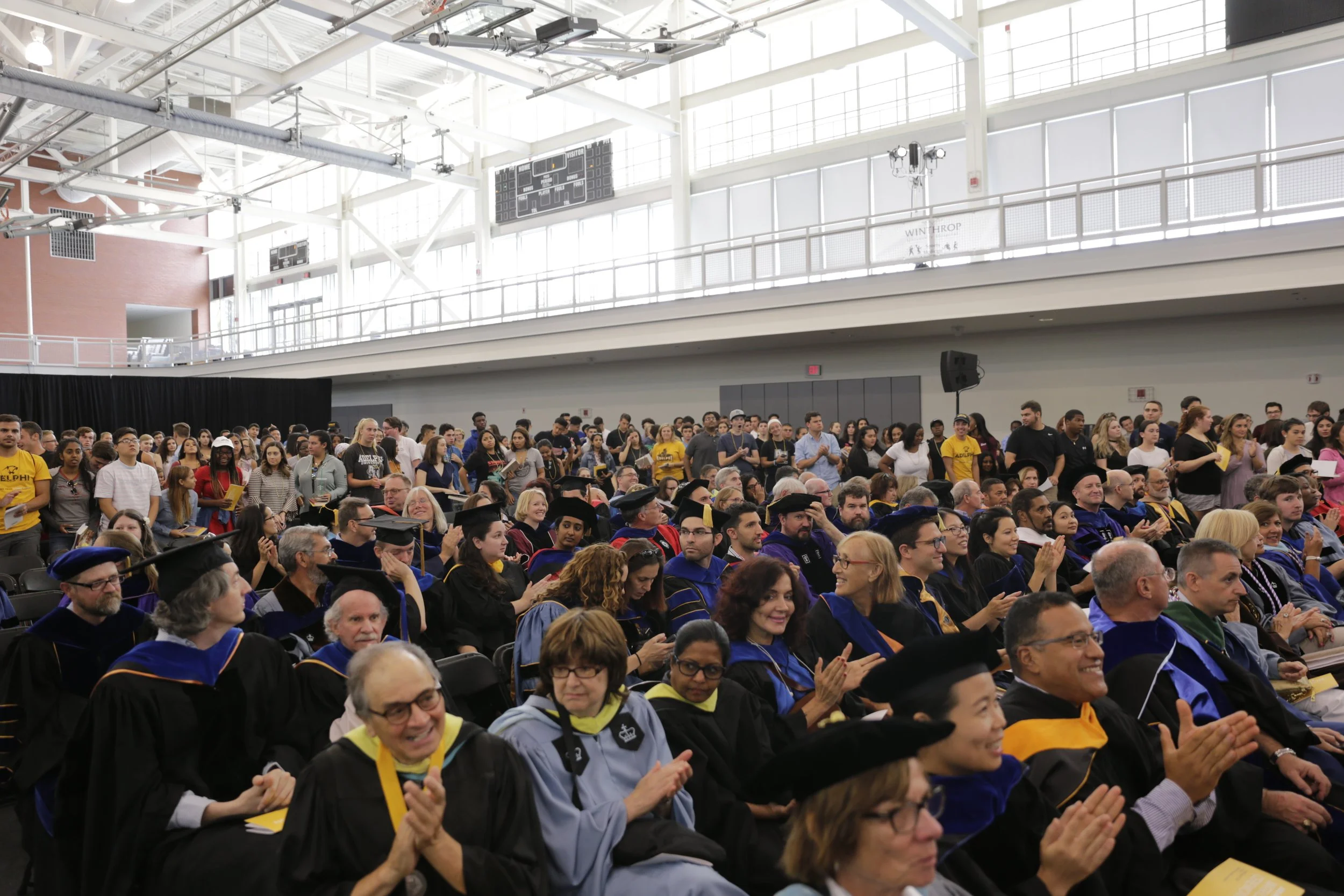 Crowd of university graduates in caps and gowns attending a commencement ceremony in a large indoor gymnasium.