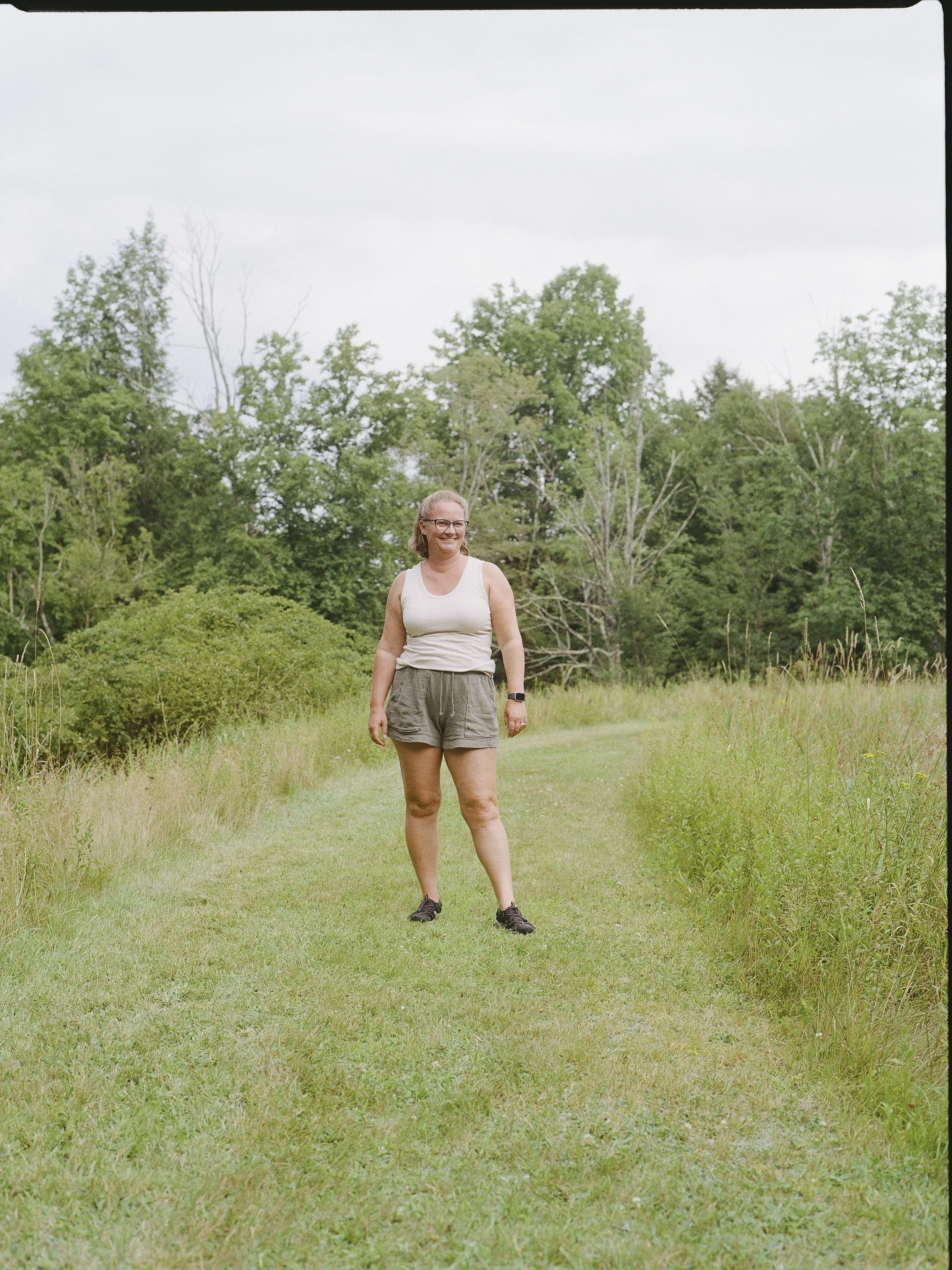 A woman standing on a grassy field with trees in the background during overcast weather. She is wearing a sleeveless top, shorts, and glasses, smiling and looking to her left.