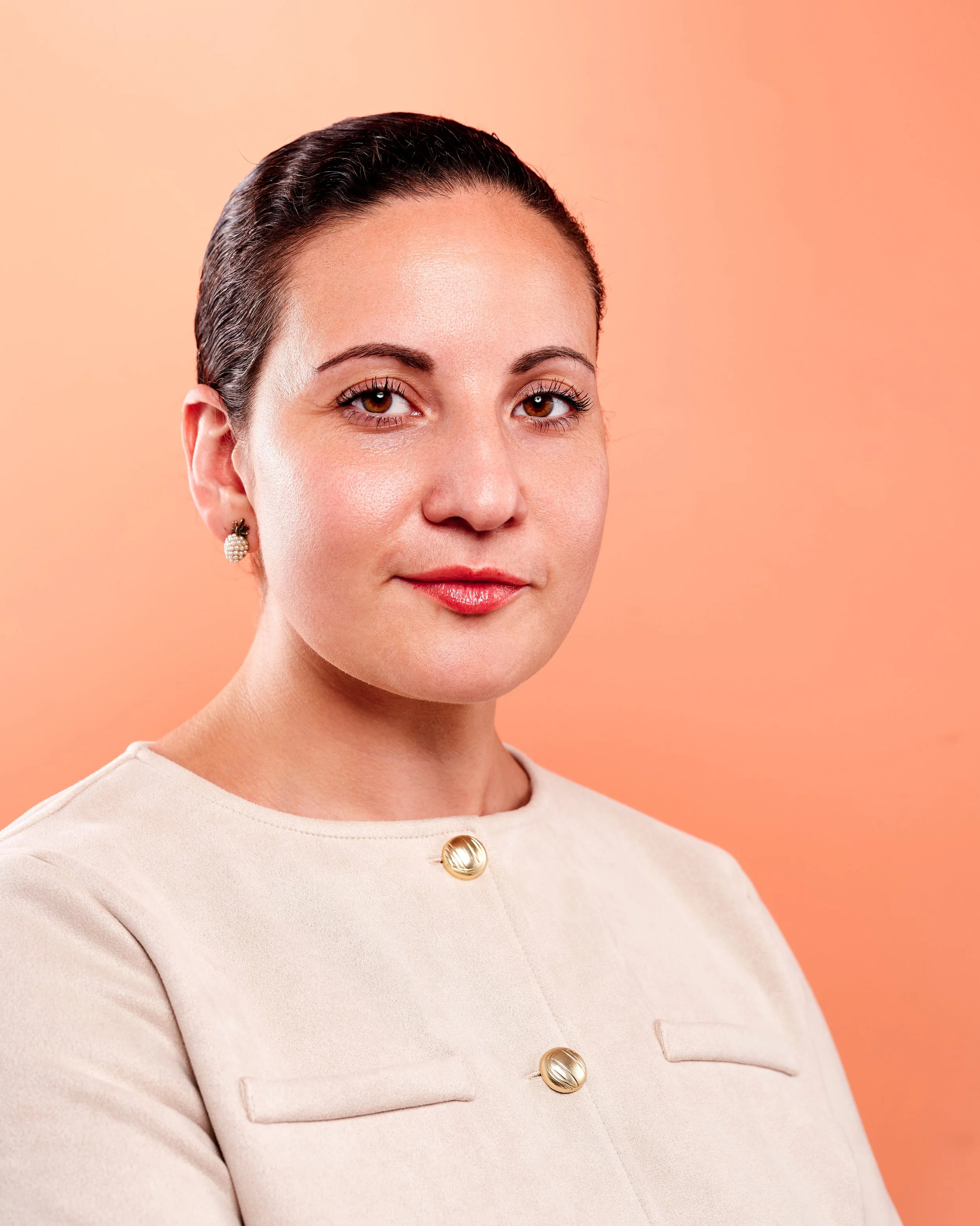 Close-up of a woman with dark hair slicked back, wearing pearl earrings and a cream-colored top with gold buttons, against a peach background.
