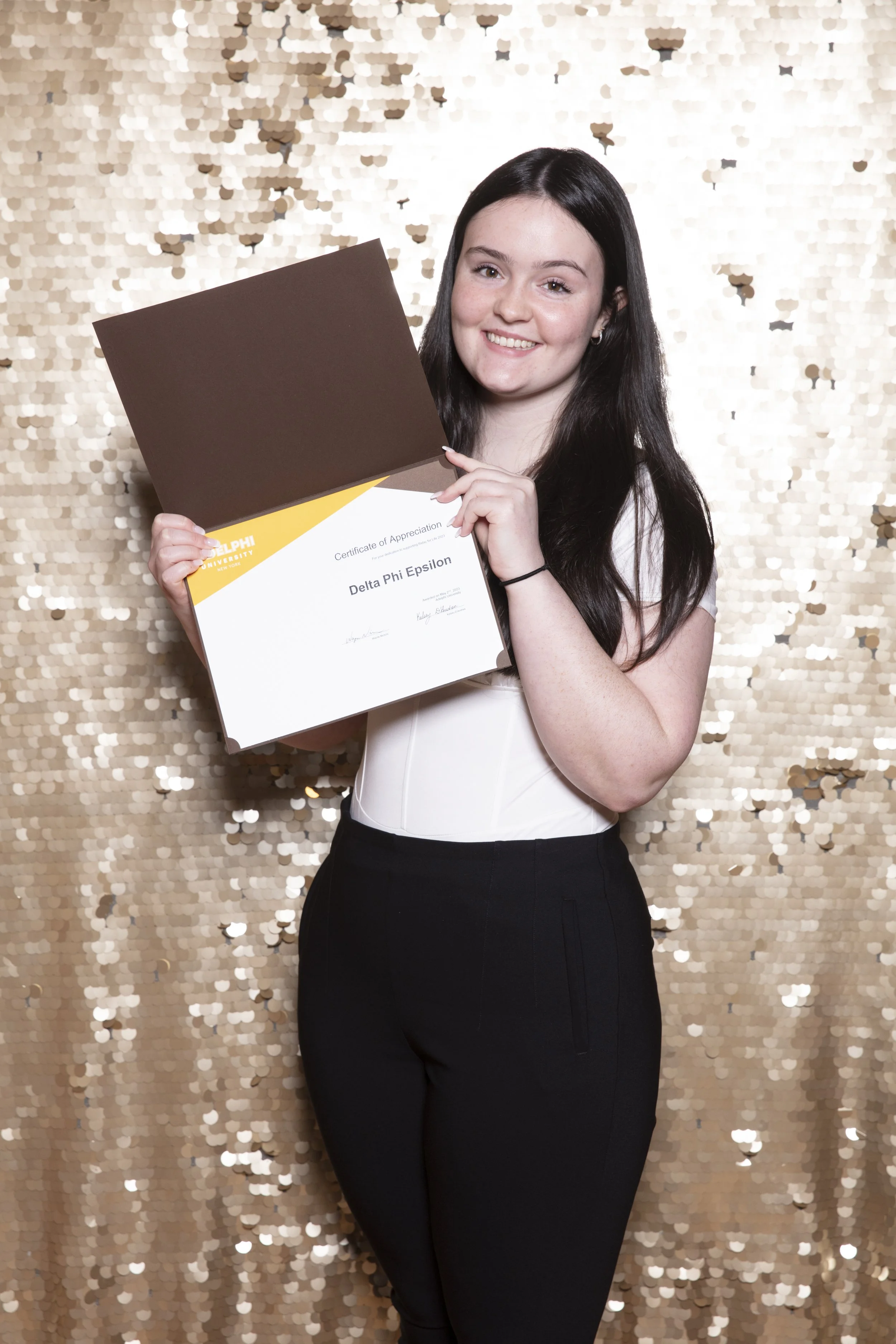 A young woman with black hair smiling and holding a diploma in front of a sparkling gold backdrop.