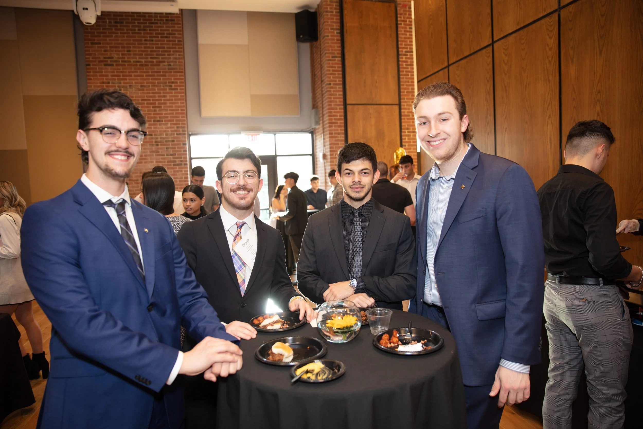 Four young men in suits standing around a table at a formal event, smiling at the camera inside a well-lit room with wooden paneling and brick walls, other guests in the background.