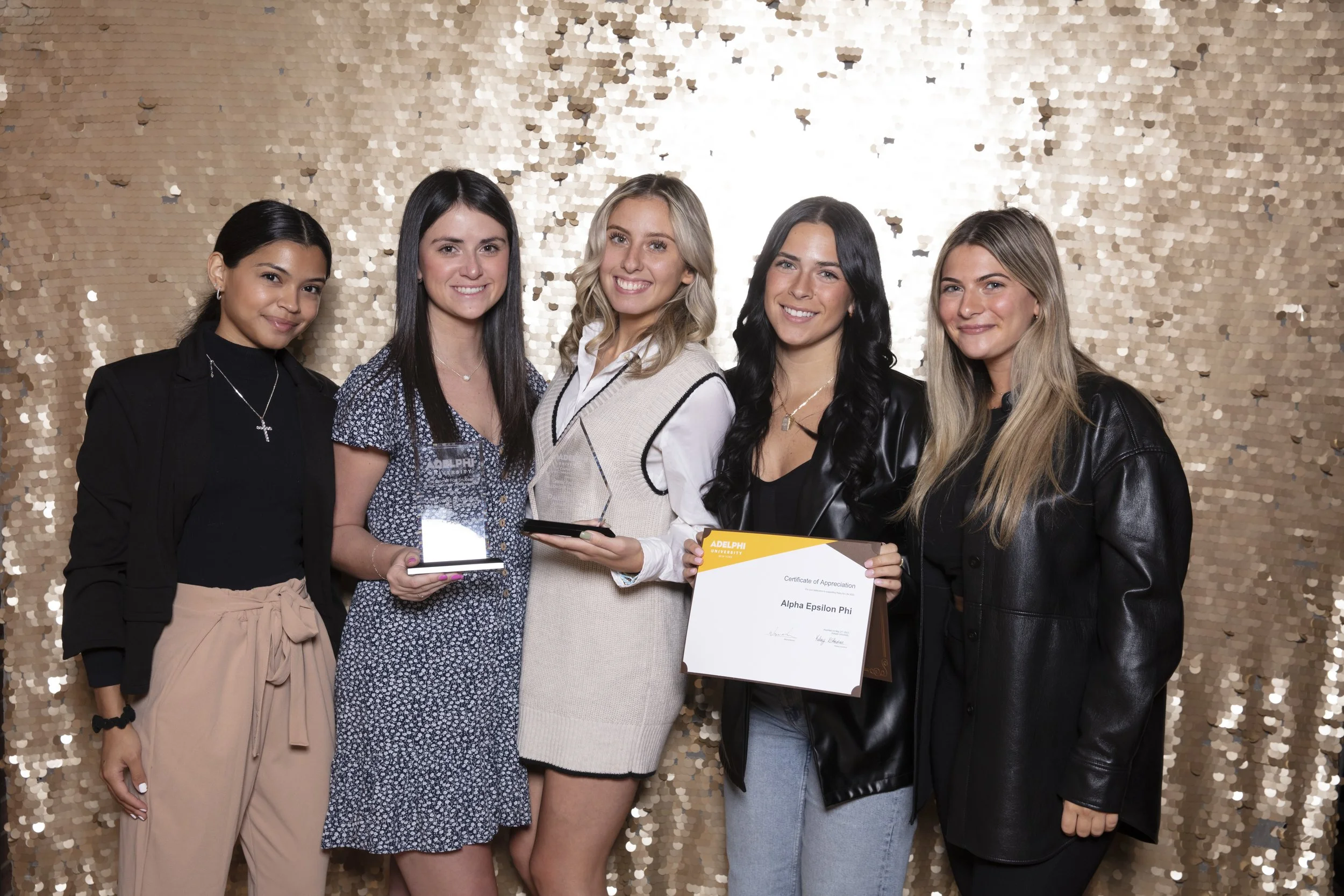 Five young women standing together, smiling, at an award ceremony in front of a gold sequin backdrop. The women are holding awards and certificates, celebrating their achievements.
