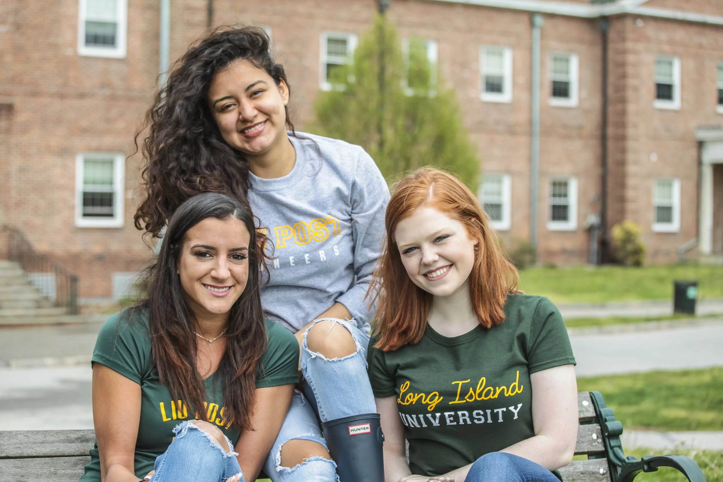 Four young women sitting on a park bench outdoors, smiling, with a brick building in the background. Two are wearing green Long Island University T-shirts and one is wearing a gray sweatshirt. They appear to be students.