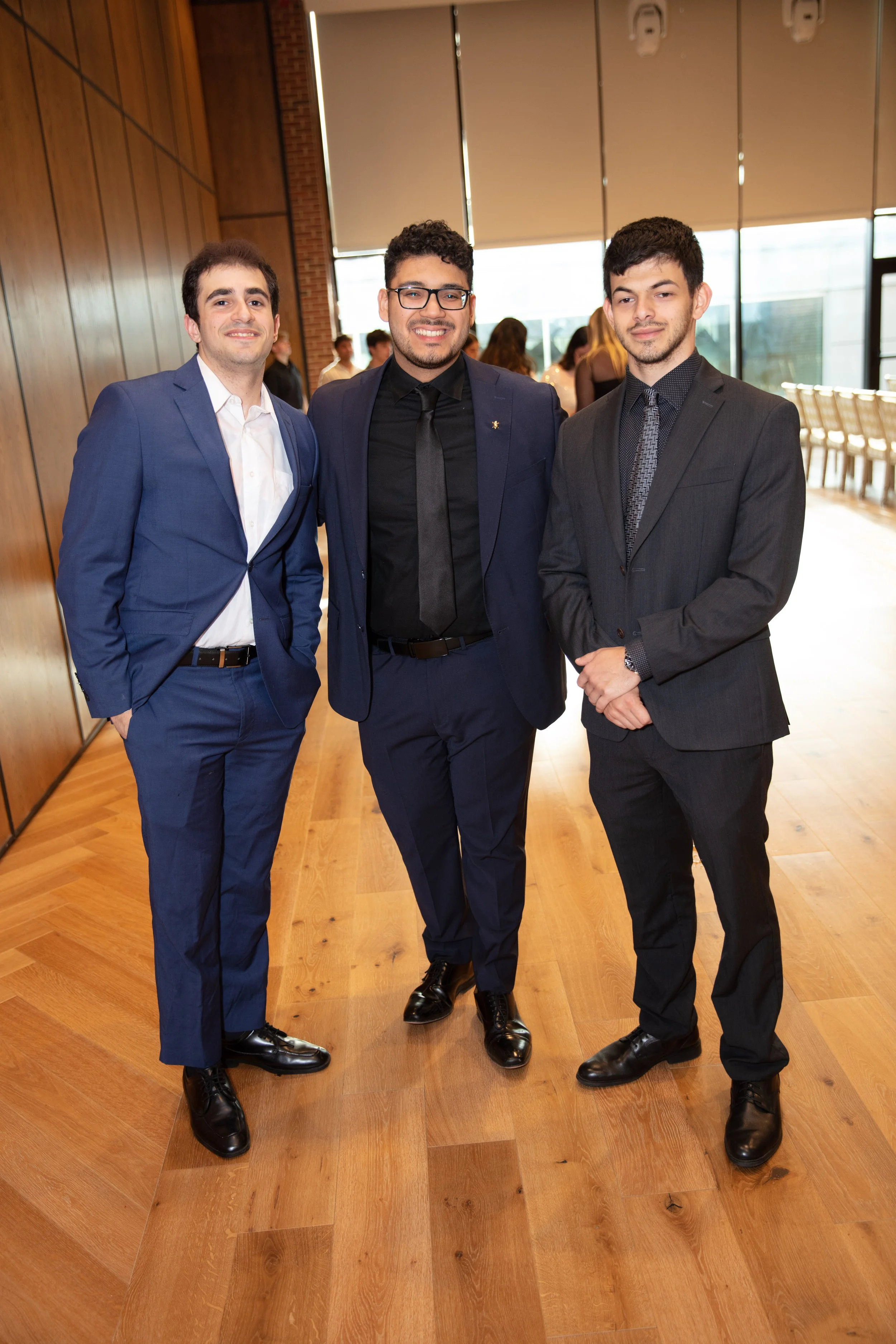 Three young men in suits standing together indoors, smiling at the camera, at a formal event or gathering.