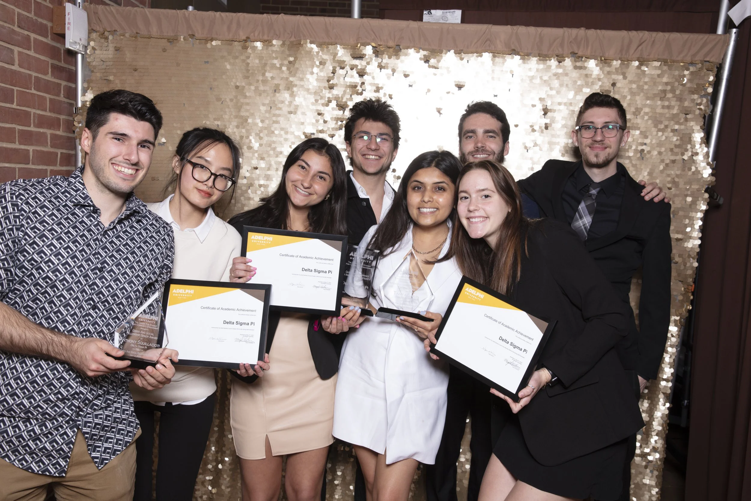 Group of nine young adults, smiling, holding certificates and awards at an award ceremony, with a gold sequin backdrop.