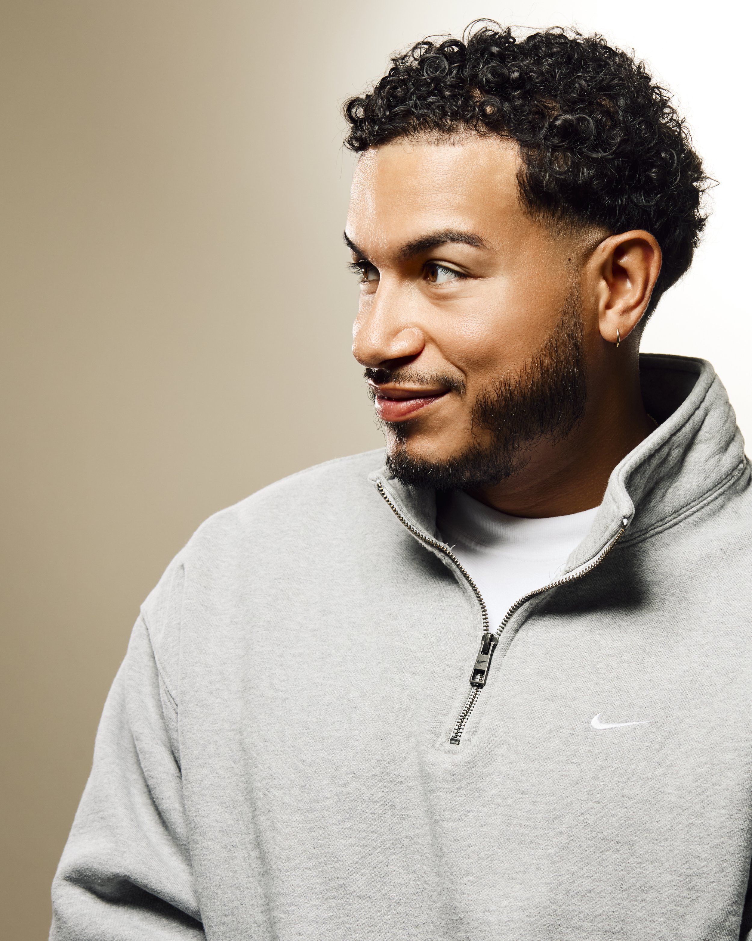 A young man with a beard and curly dark hair, wearing a gray hoodie with a small Nike logo, smiling and looking to his left, against a neutral background.