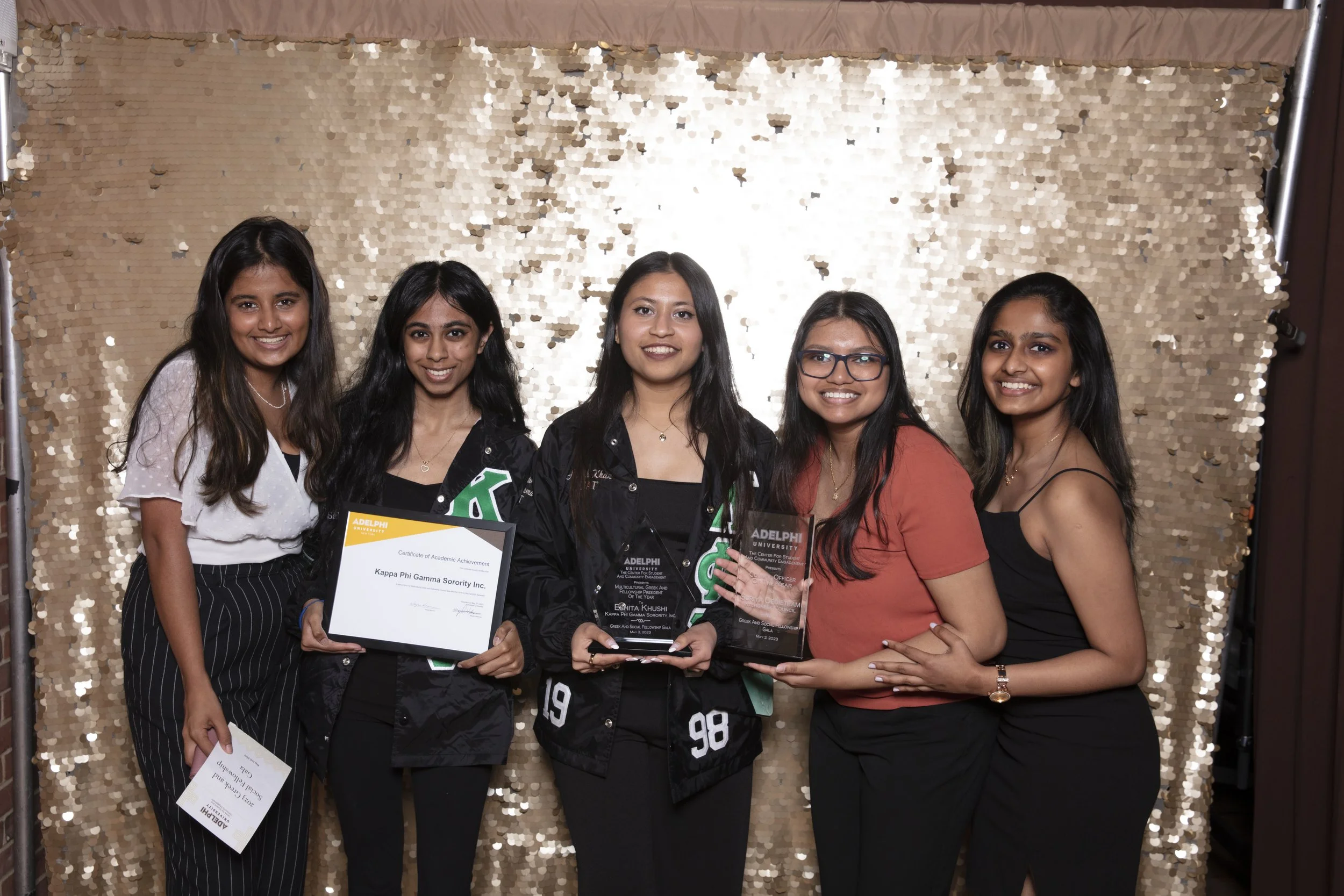 Five women standing together, smiling, holding awards and a certificate, in front of a shiny gold sequin backdrop.