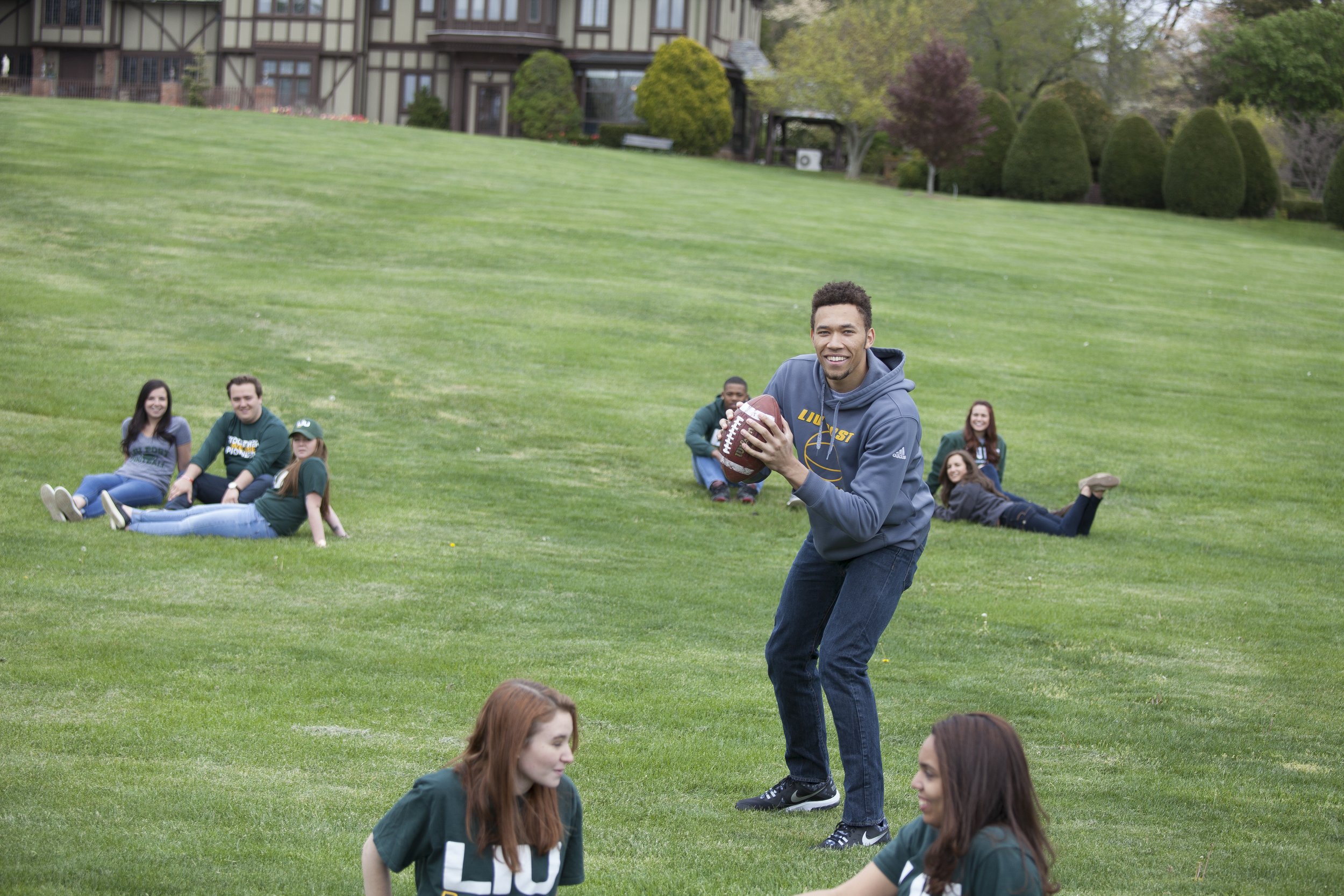 A young man in a gray hoodie holding a football on a grassy hill, with several people sitting and lying on the grass in the background, and a house with trees in the distance.