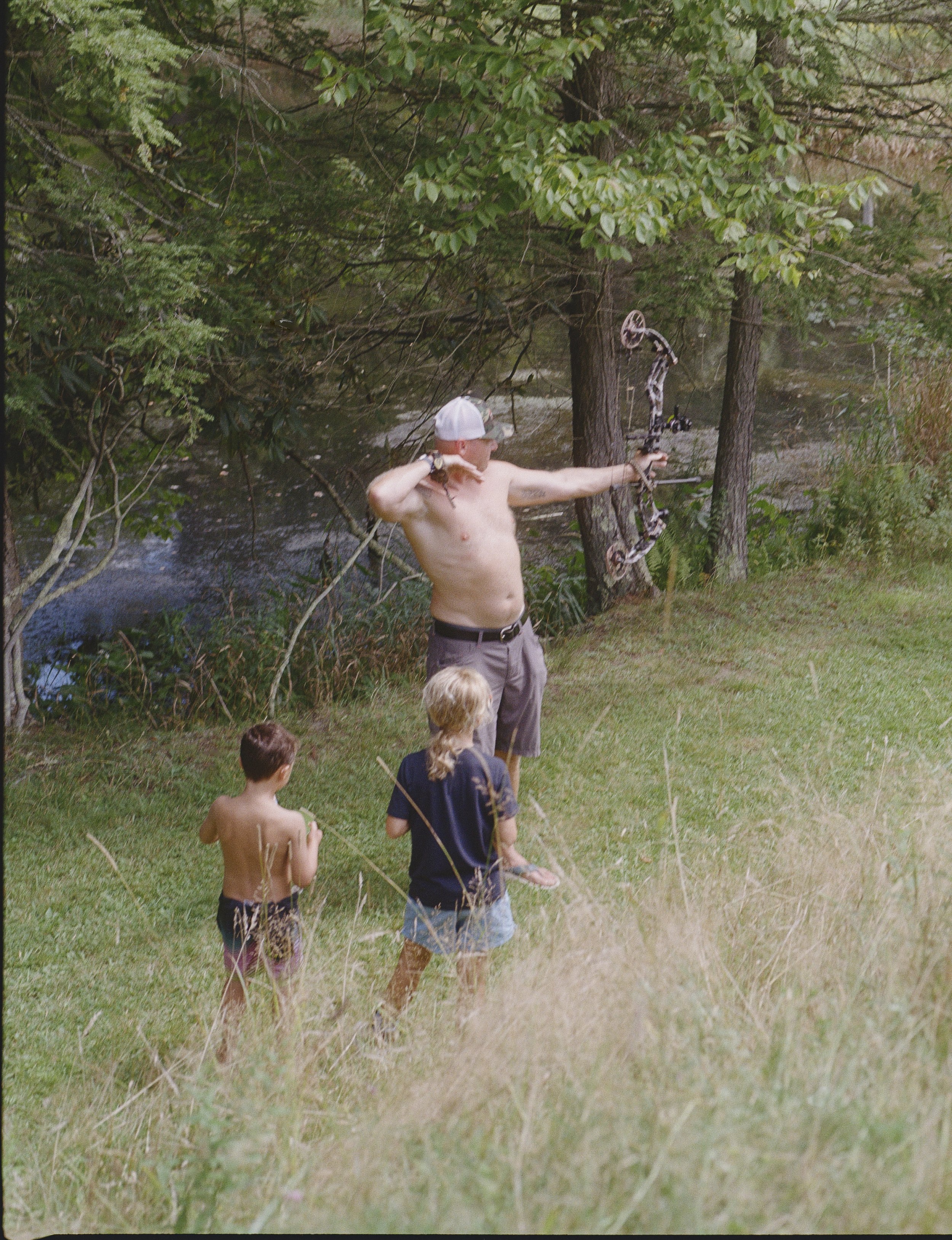 A man holding a bow and arrow standing outdoors near a river, with three children watching him. The man is shirtless, wearing a baseball cap, and appears to be aiming the bow. Trees and grass surround the scene.