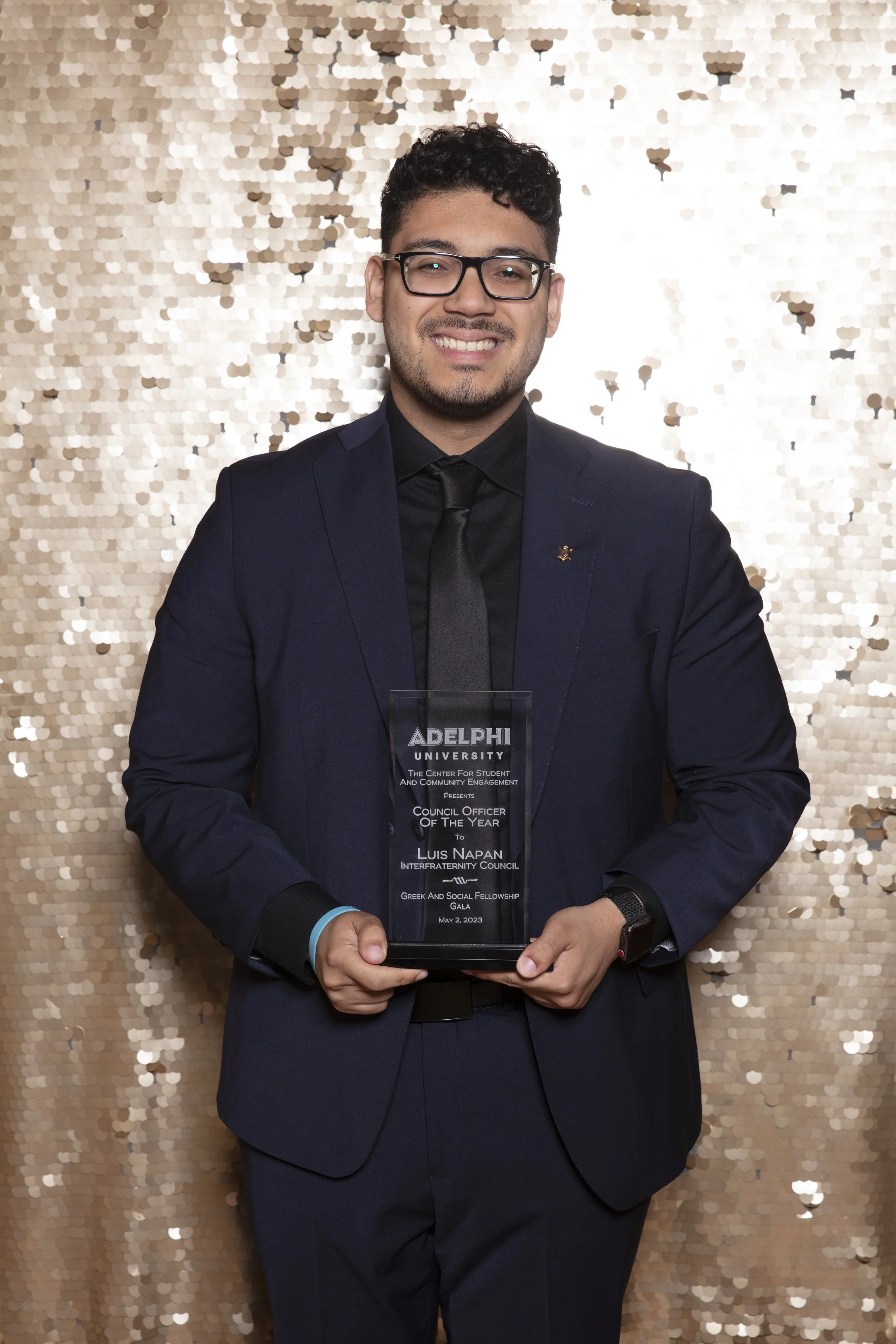 Man in navy suit and glasses holding an award plaque at an event with a gold sequin backdrop.