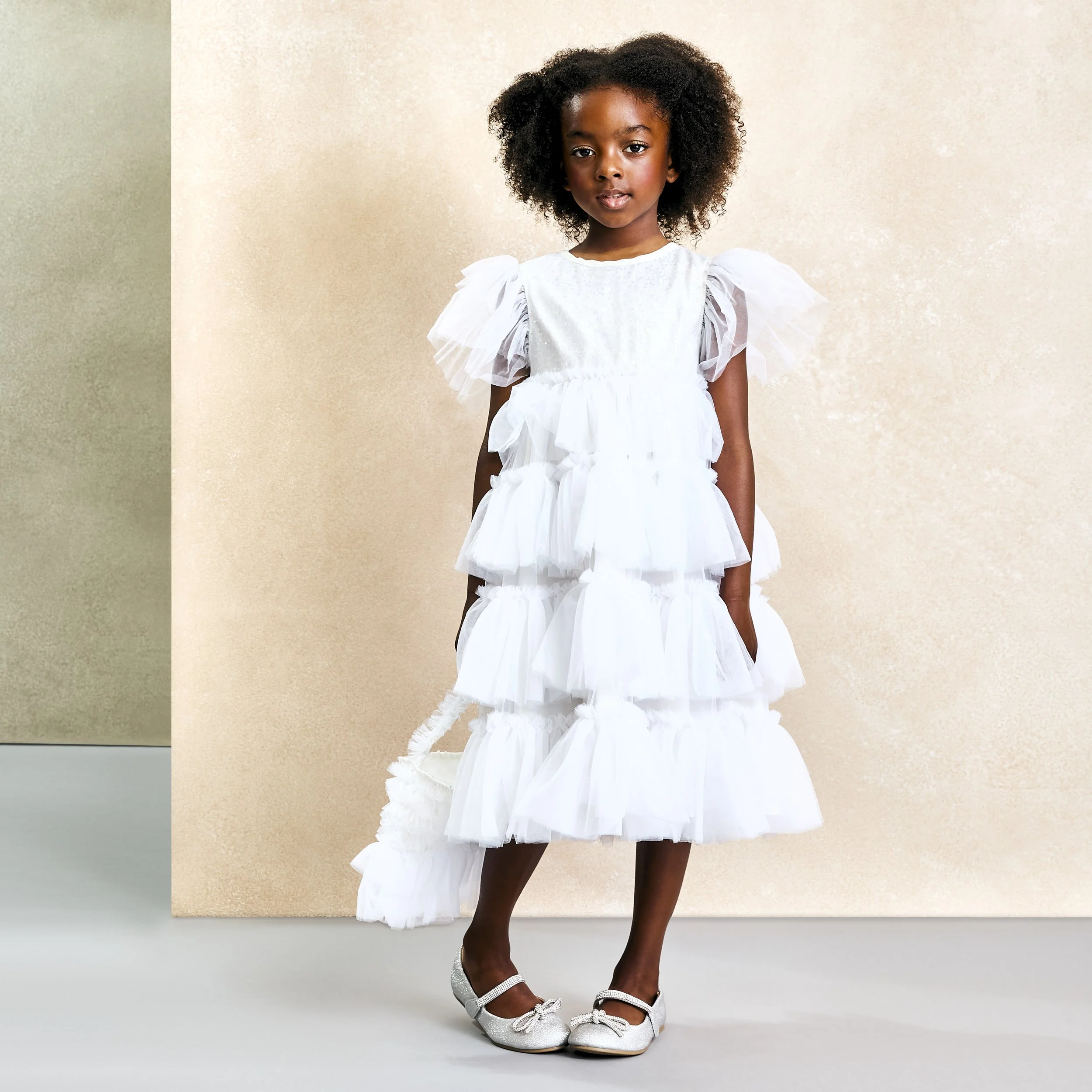 Young girl in a white, tiered dress with puffed sleeves, holding a small white bag, wearing silver shoes, standing against a neutral beige background.