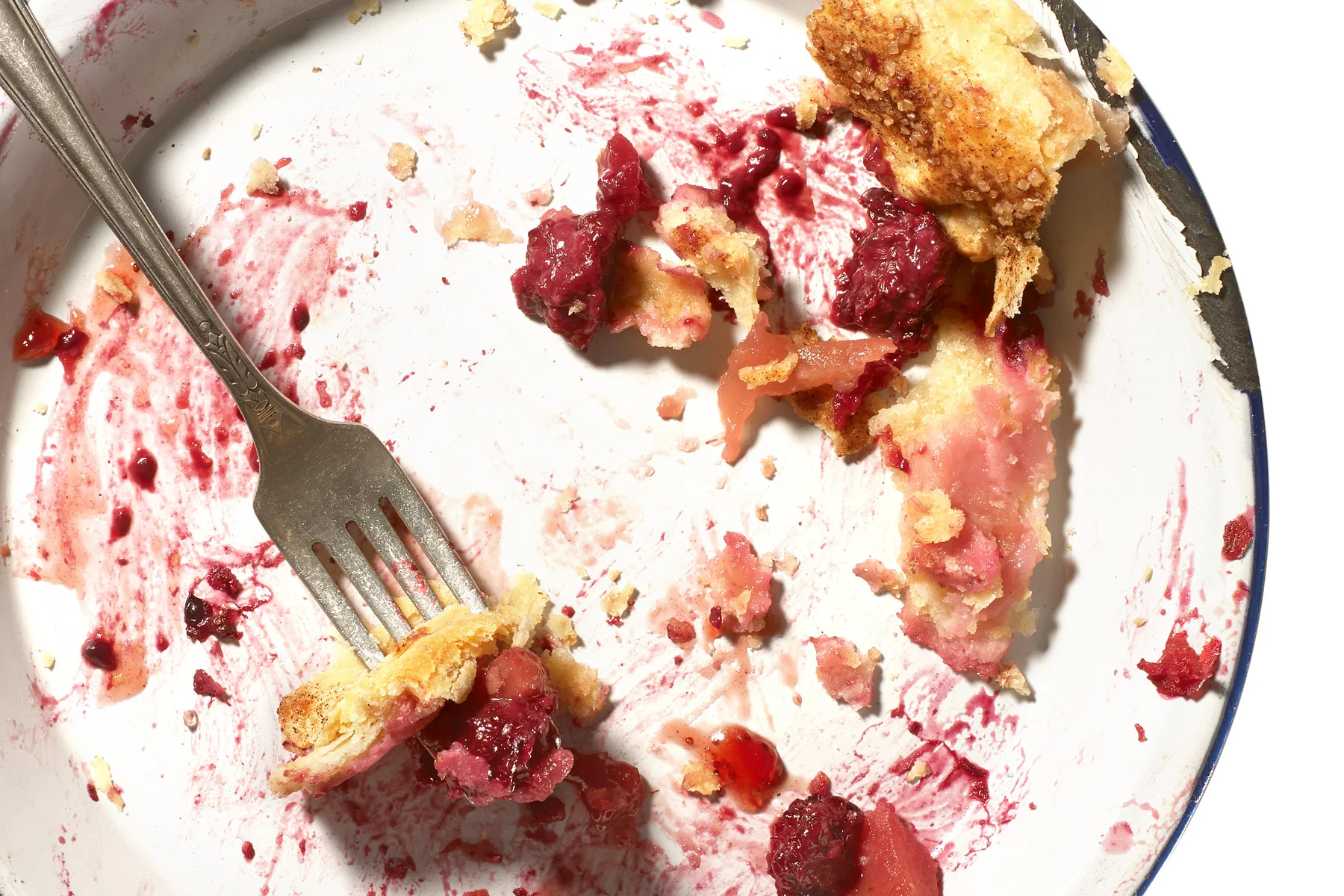 A mostly empty white plate with a blue rim, with remnants of a berry pie, including crumbs, filling, and crust pieces, and a fork resting on the plate.