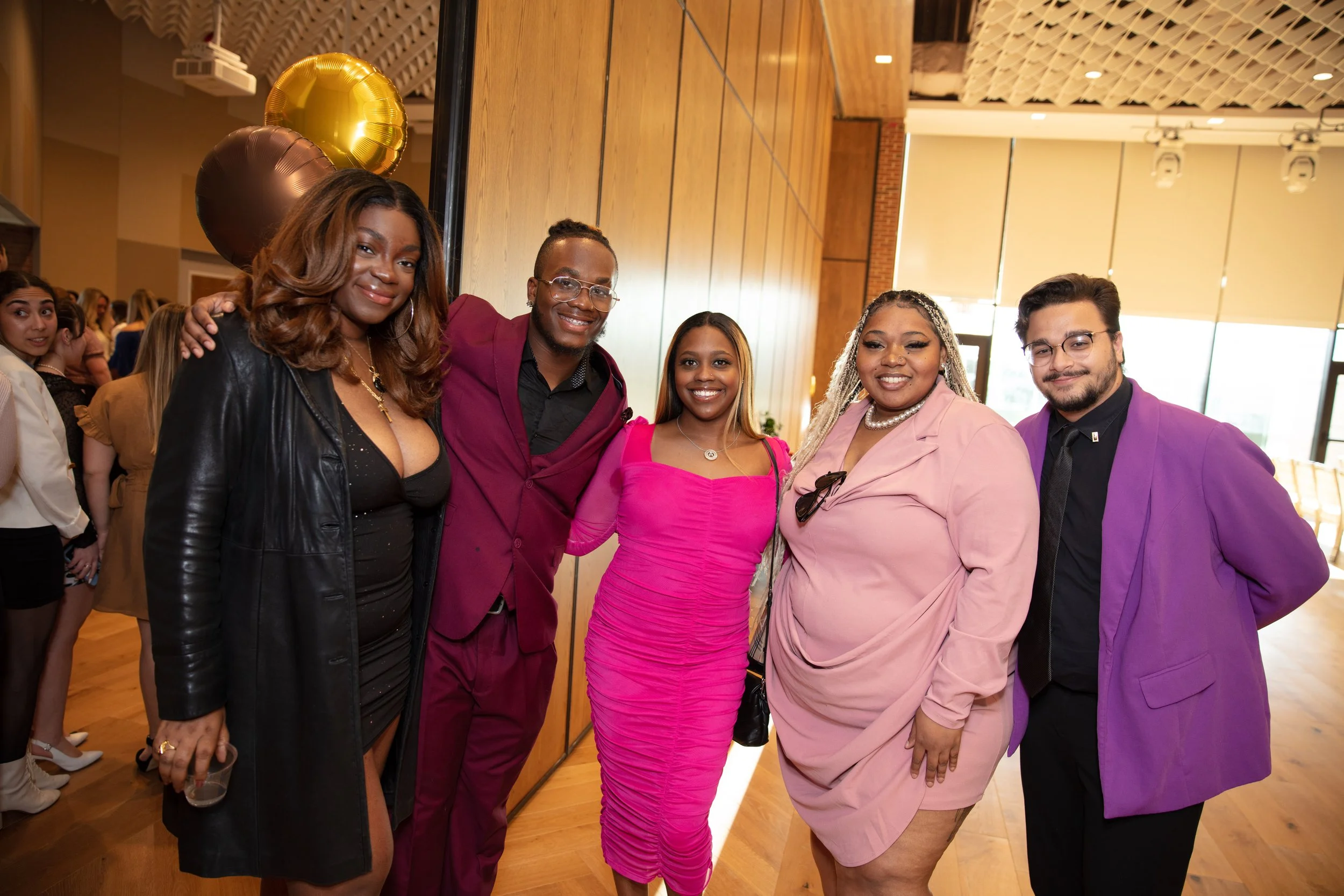 Group of five people, three women and two men, dressed in colorful formal attire, smiling and posing for a photo at an indoor event with wooden walls and large windows.