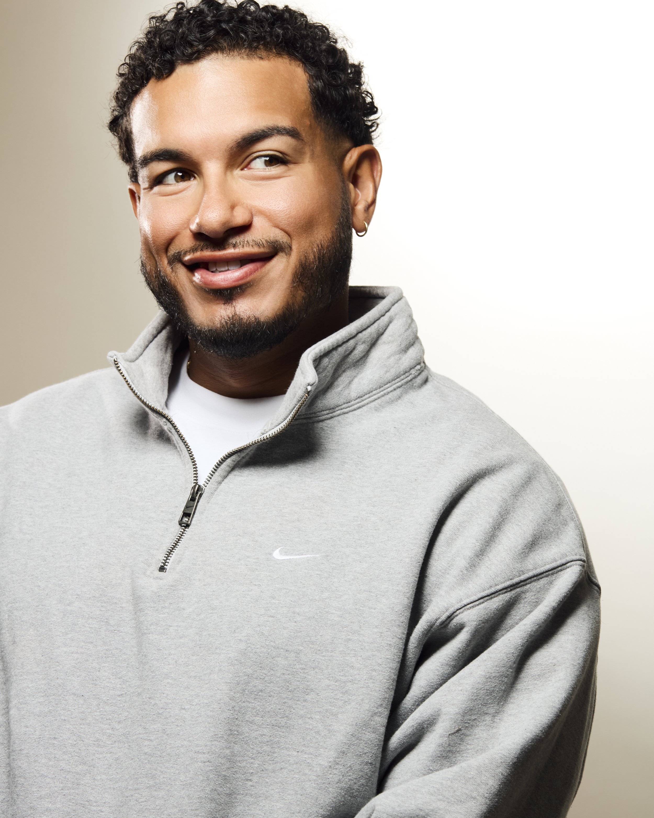 Close-up of a smiling young man with curly dark hair, facial hair, earrings, and a grey Nike hoodie against a neutral background.