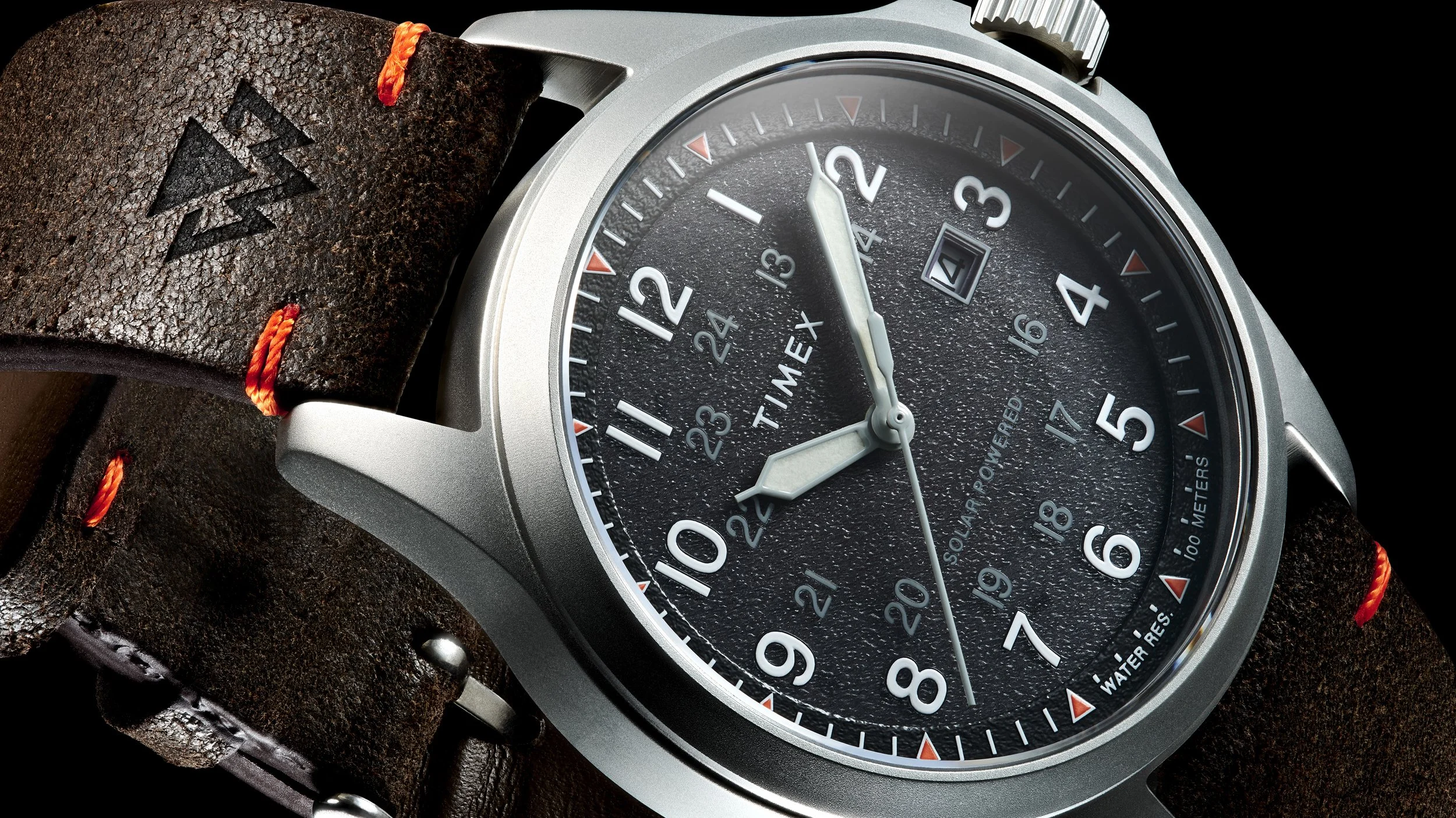 Close-up of a Timex wristwatch with a black textured dial, white numerals, and a brown leather strap with red stitching.