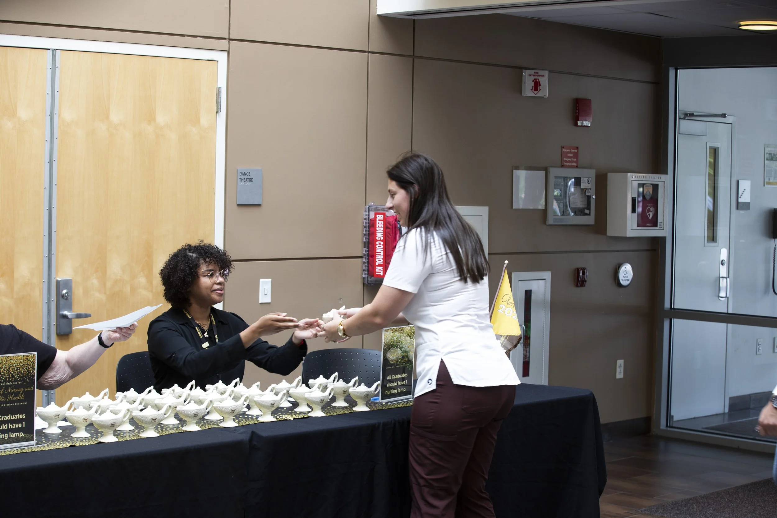 A woman in a white shirt is receiving a small item from a woman in a black shirt behind a table with many ceramic bowls at a graduation event.