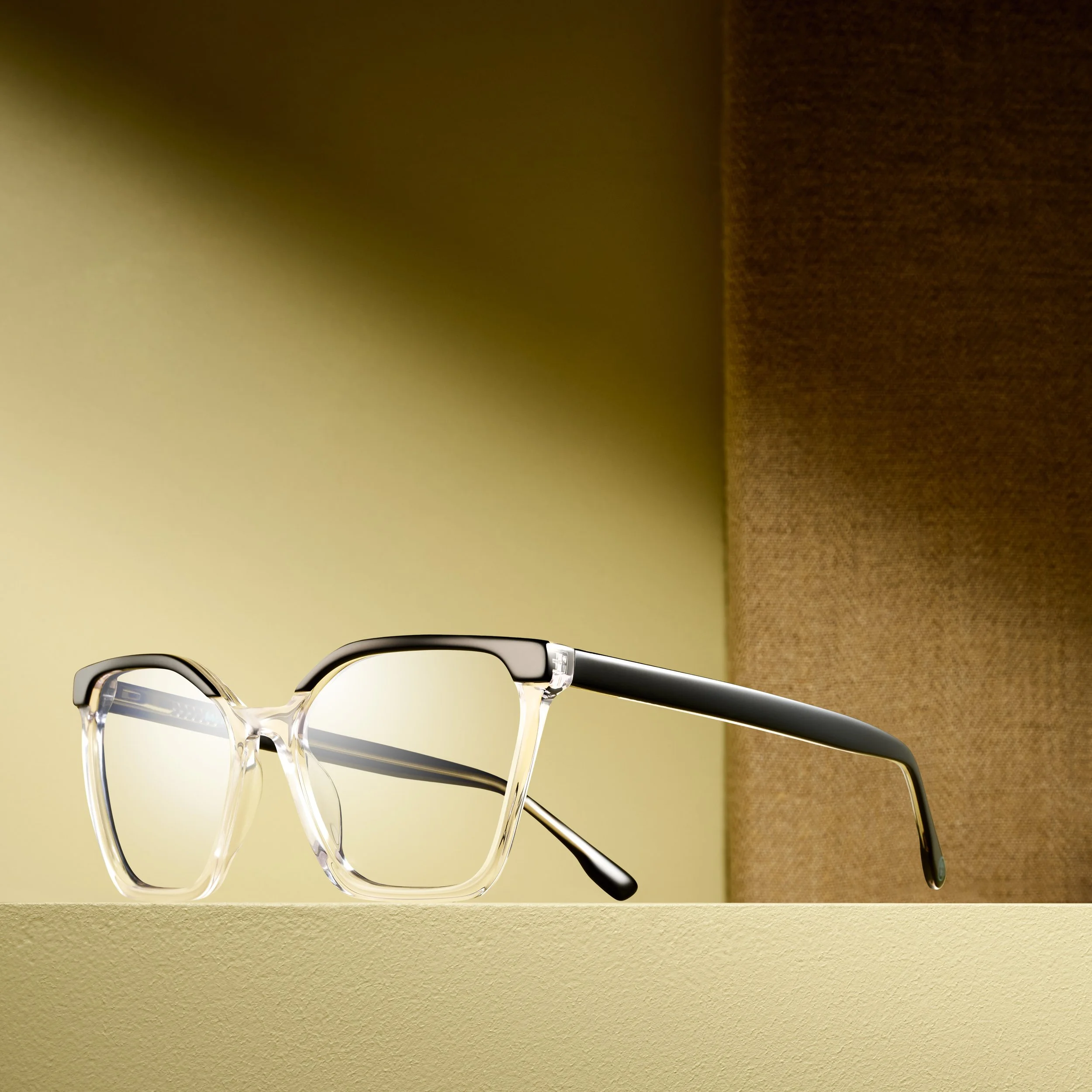 Close-up of stylish eyeglasses with clear frame and black temple rests on a beige surface with a background featuring a gradient from light yellow to dark brown