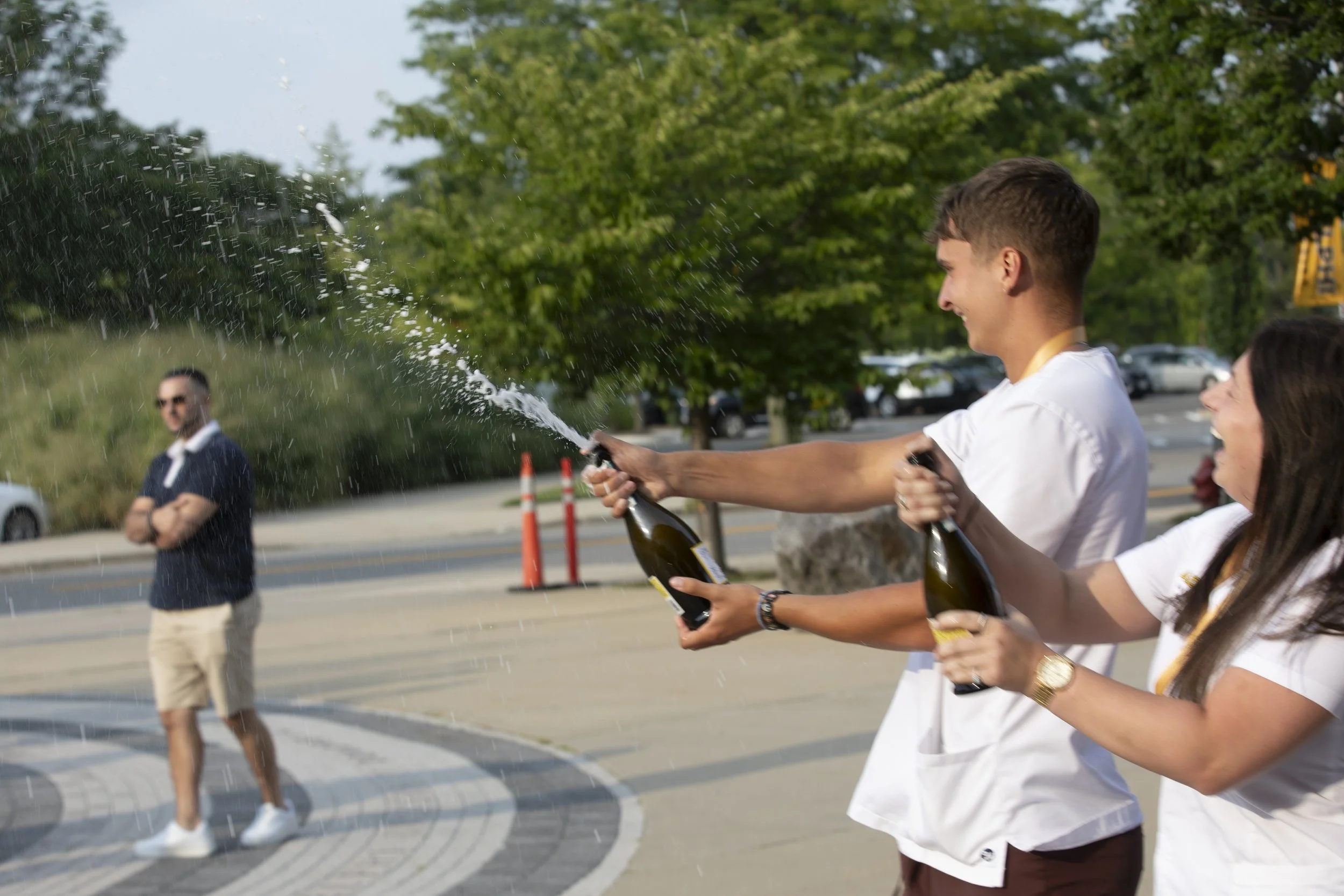 Two young people celebrating outdoors by opening champagne bottles and spraying champagne towards a man standing nearby on a sidewalk with parked cars and trees in the background.