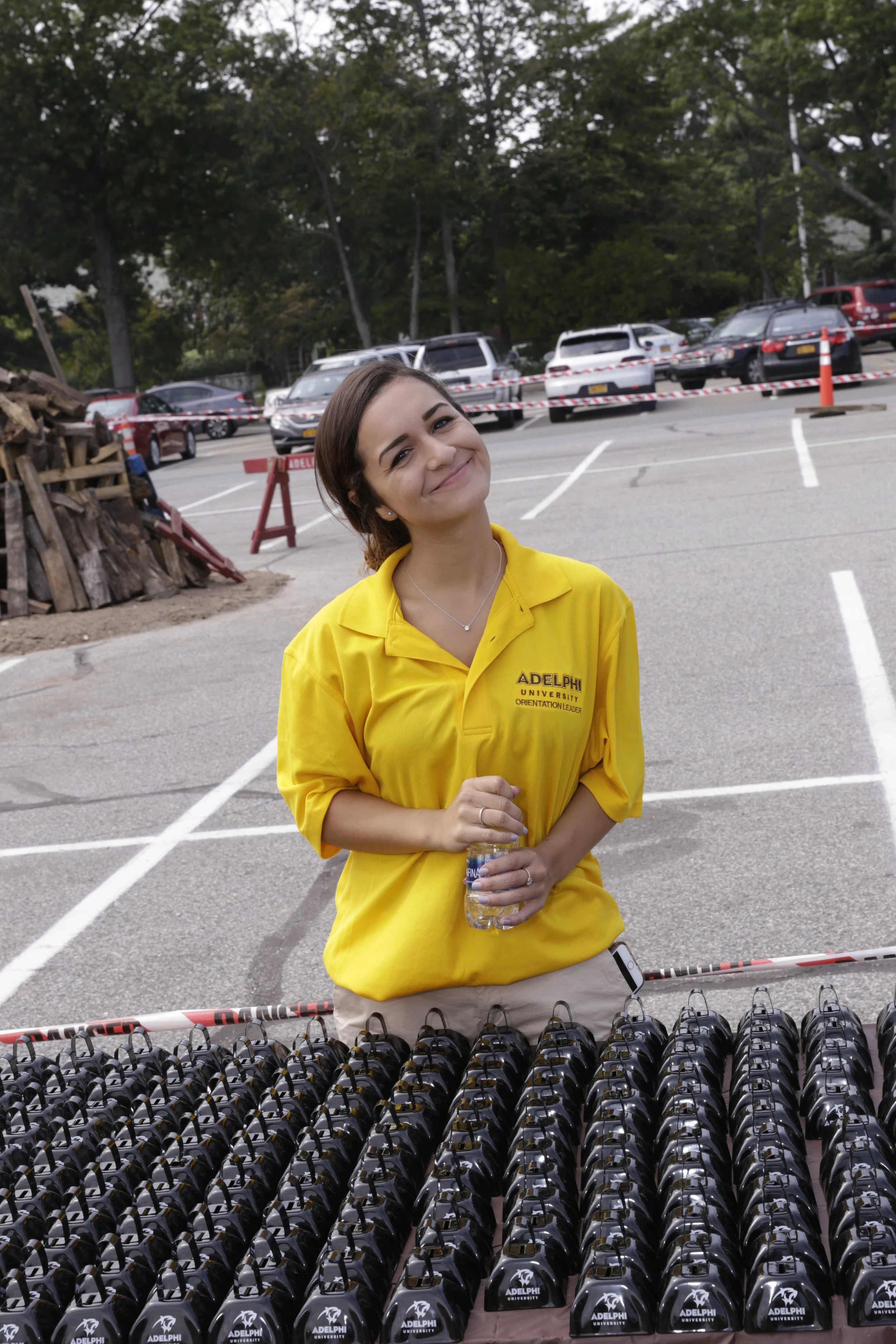 A young woman in a yellow Adelphi University polo shirt stands behind sunglasses arranged on a table, holding a water bottle, smiling at the camera outdoors in a parking lot.
