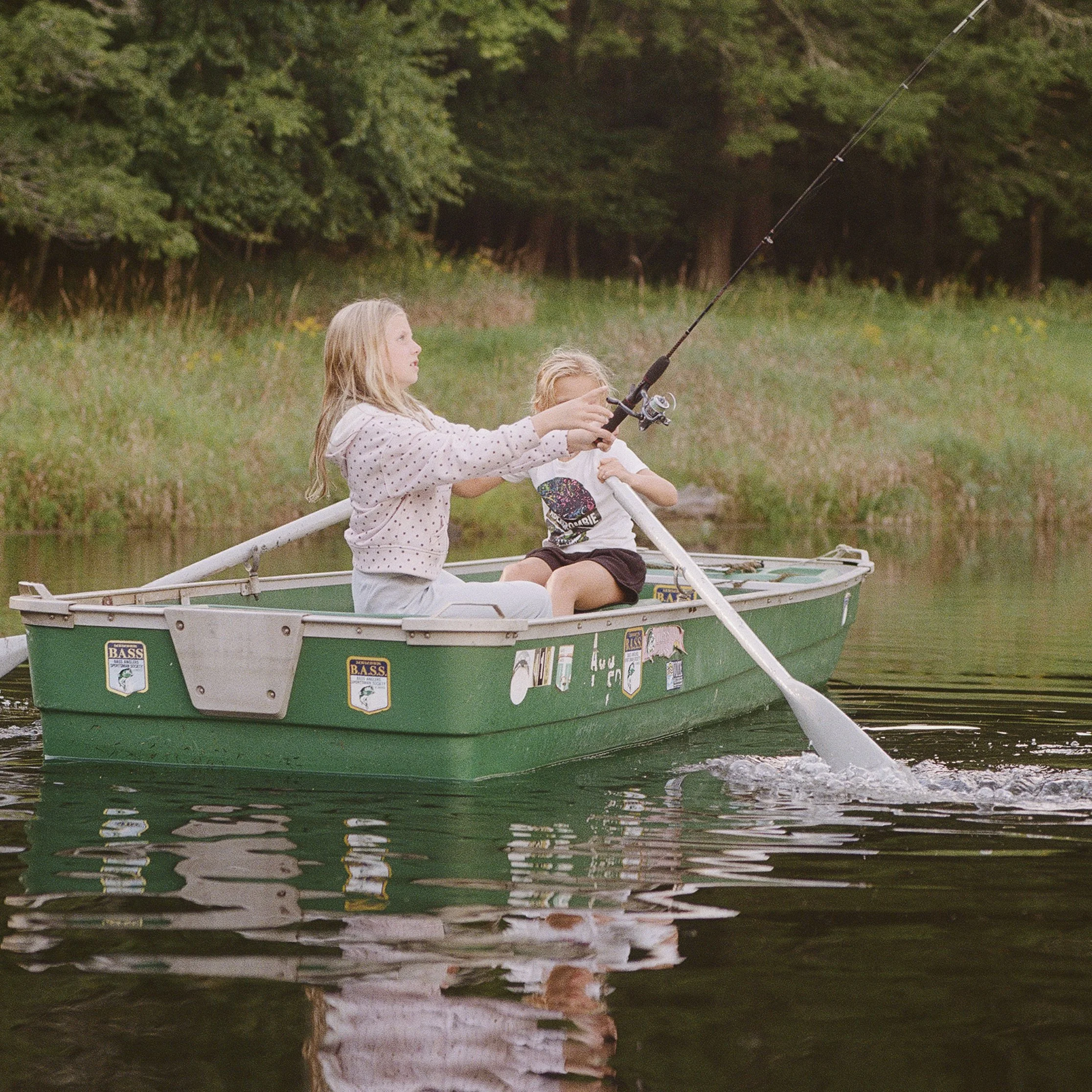 Two young girls fishing from a small green boat on a calm lake surrounded by trees.