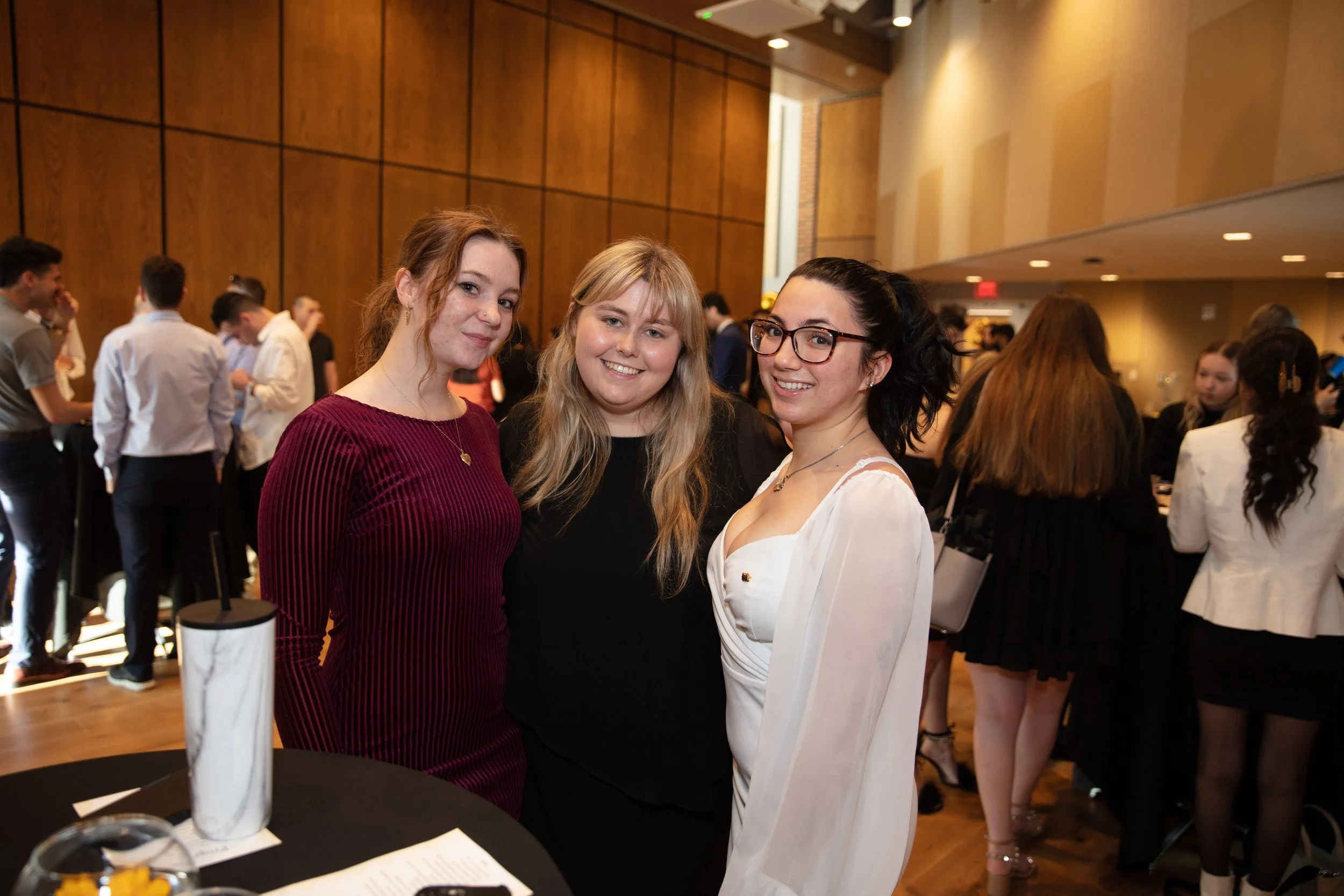 Three young women posing together at a social event in a large, well-lit room with wooden wall panels, surrounded by other attendees.