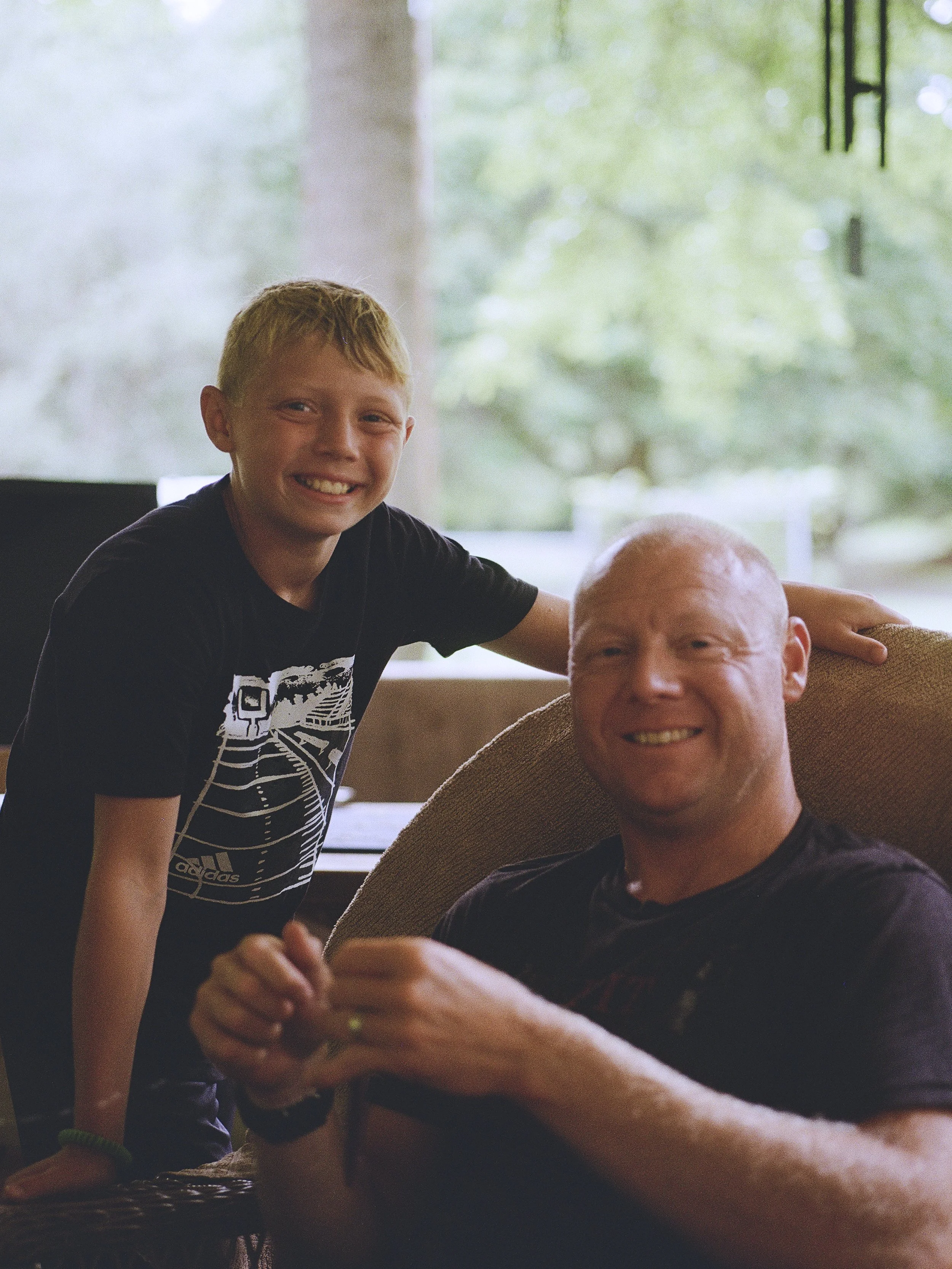 A young boy with blonde hair smiling, leaning on what appears to be an older man's head, in a room with a large window and trees outside.
