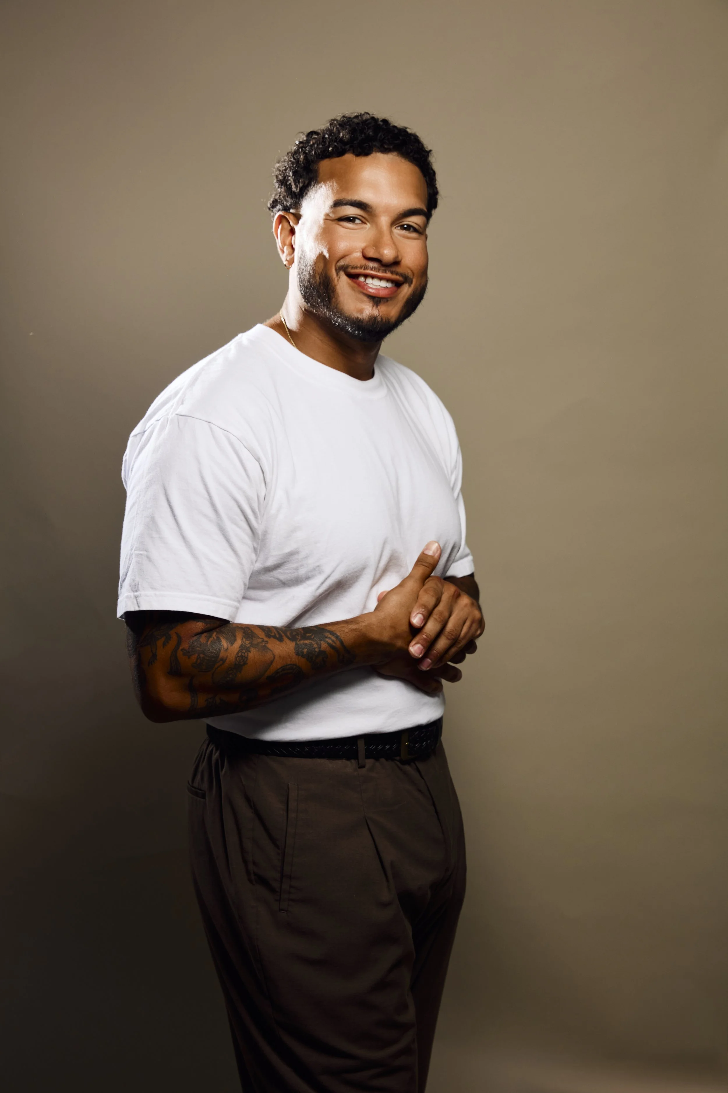 A smiling man with curly hair and a beard, wearing a white t-shirt and brown pants, standing against a plain beige background.