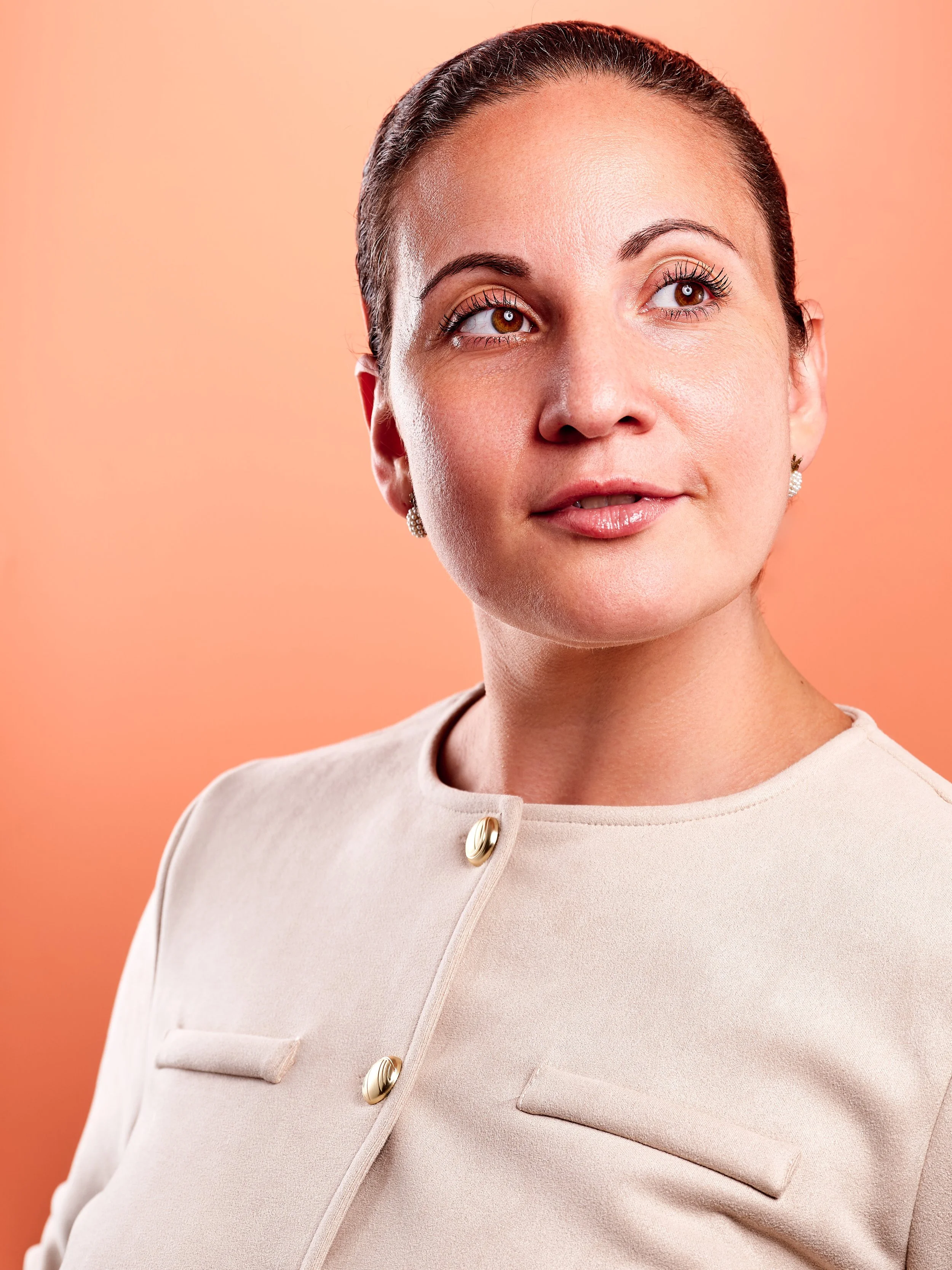 Close-up of a woman with brown hair styled in a sleek bun, wearing earrings, and a beige top with gold buttons, against a peach-colored background.
