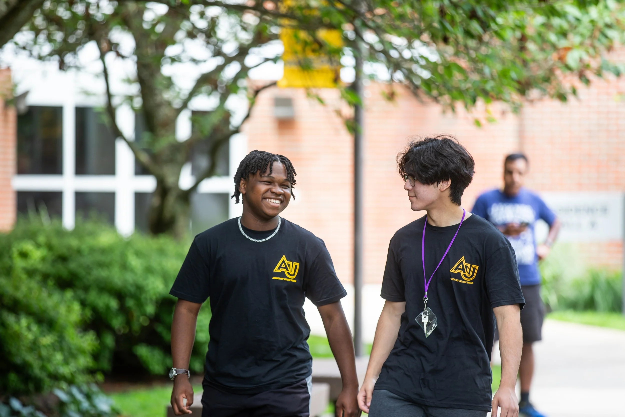 Two young men wearing black shirts with yellow 'AU' logo walking and talking outdoors, smiling at each other, with trees and a building in the background.