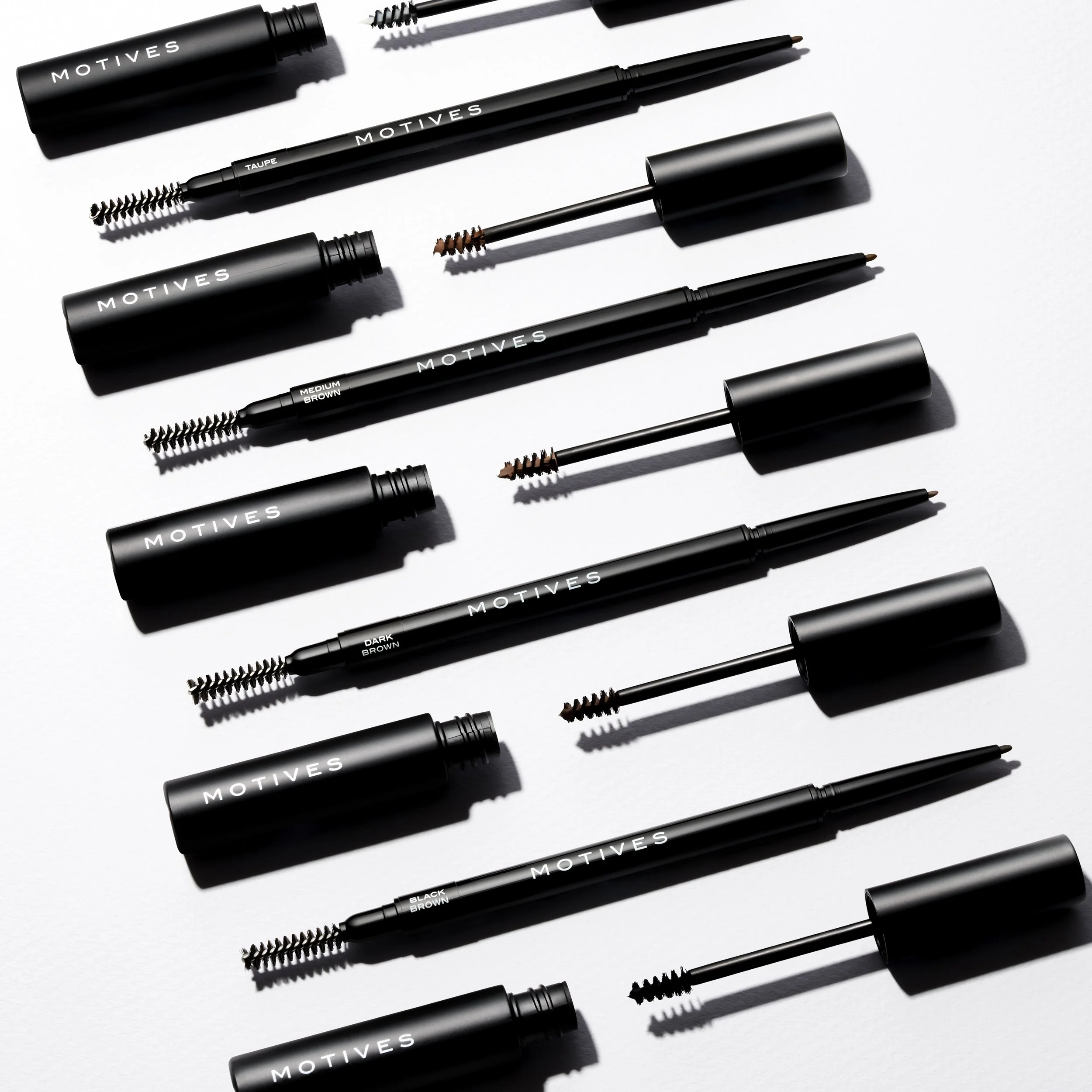 Set of black cosmetic tools with white 'Motives' branding, including mascaras, eyebrow gels, and pencils arranged on a white surface.
