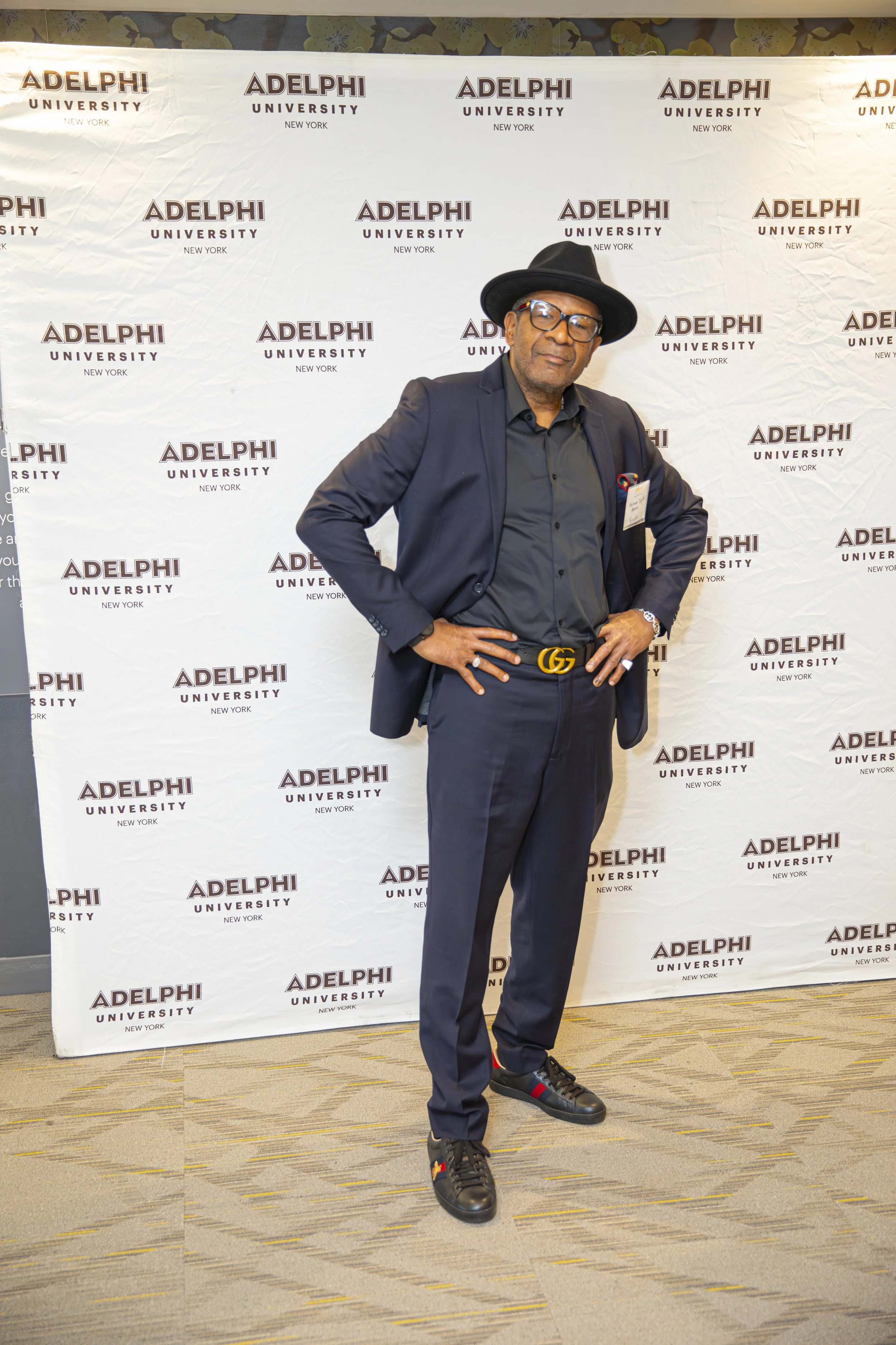 A man in a dark suit, black hat, and glasses stands with hands on hips in front of a backdrop with 'Adelphi University New York' logos.