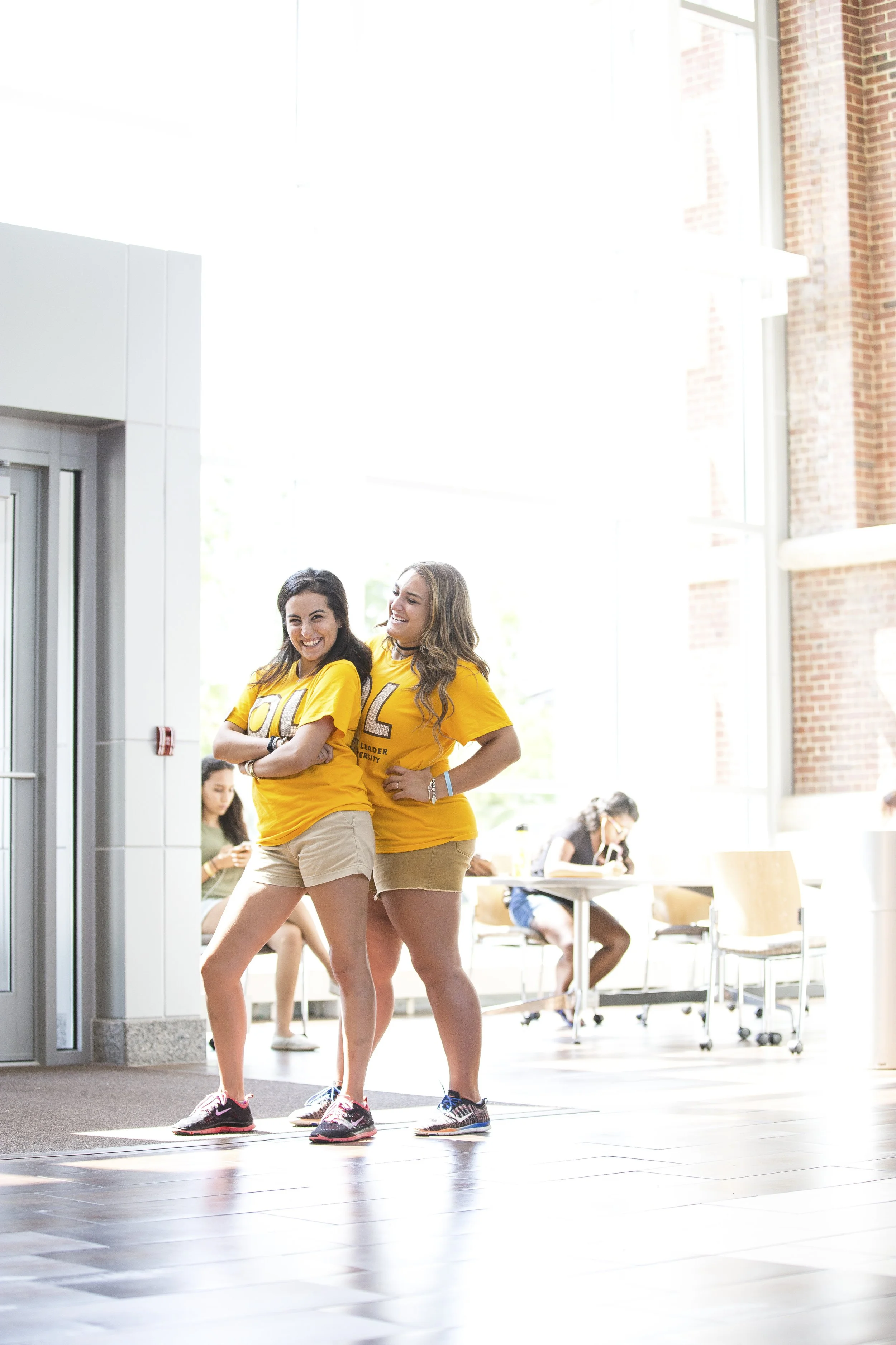 Two young women in yellow T-shirts and shorts smiling and standing together in a brightly lit indoor space with large windows, chairs, and other people in the background.