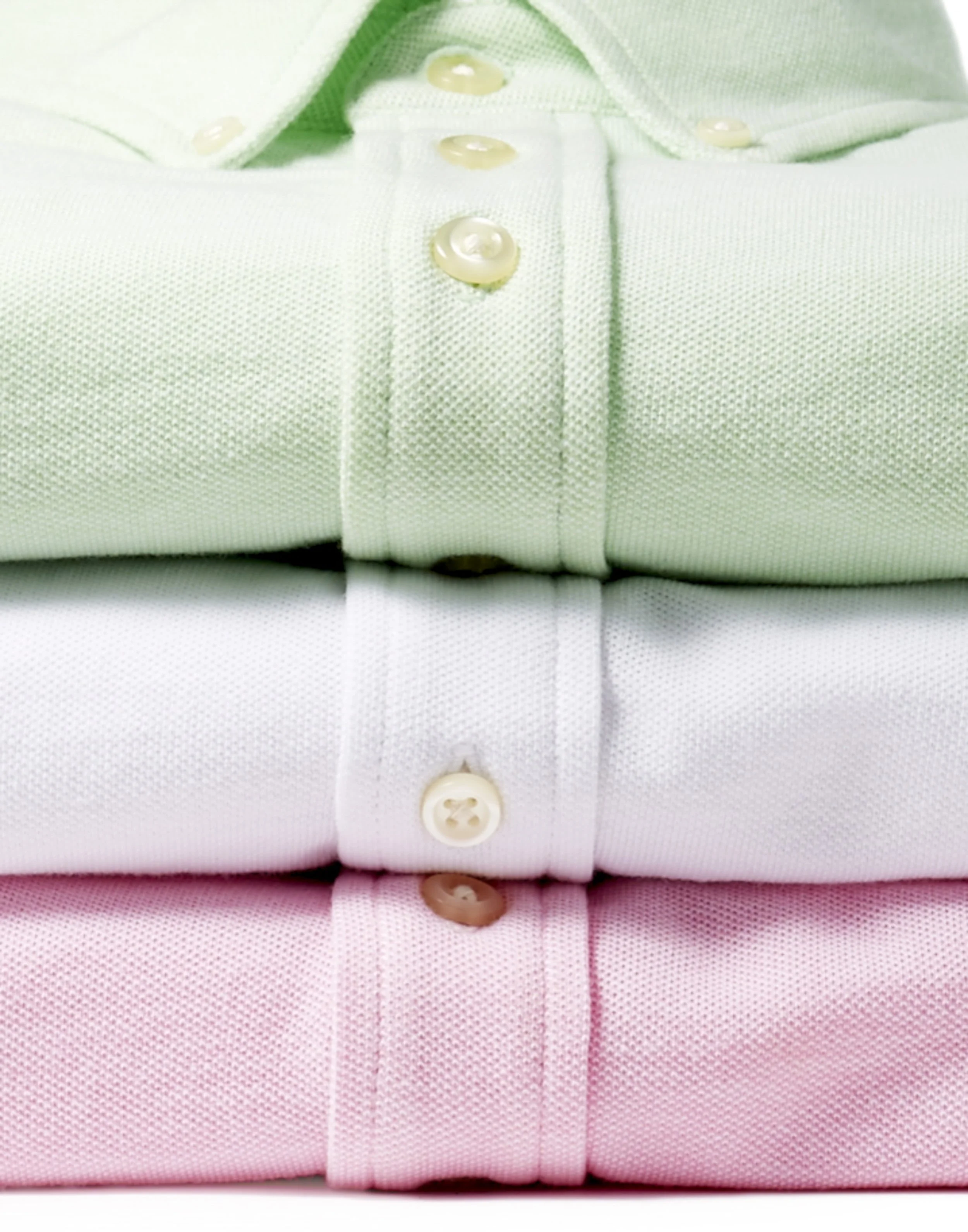 Close-up of three folded dress shirts in pastel colors: green, white, and pink, with visible buttons with two holes.
