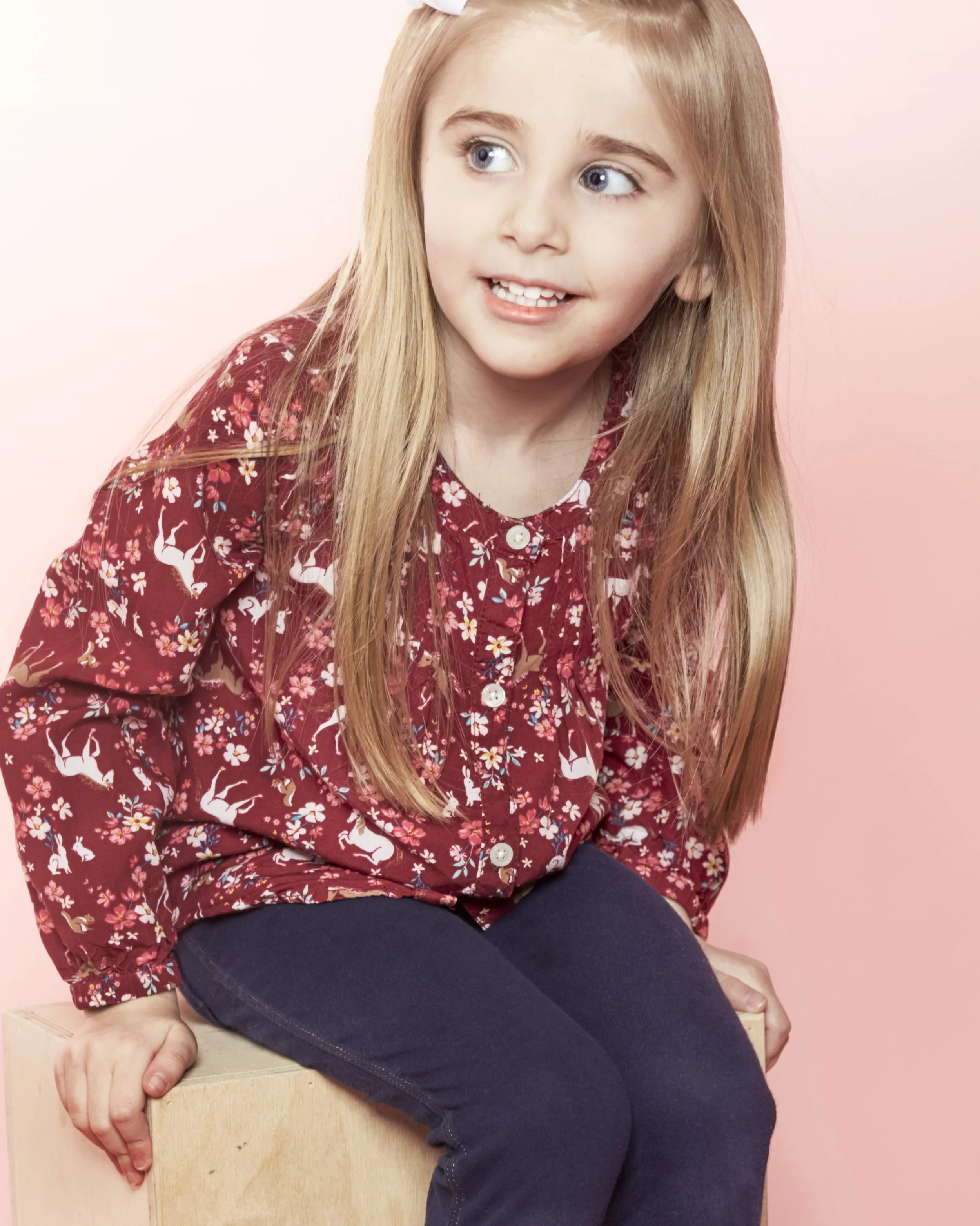 A young girl with long blonde hair and blue eyes sitting on a wooden block, wearing a red floral shirt and dark blue jeans, against a pink background.