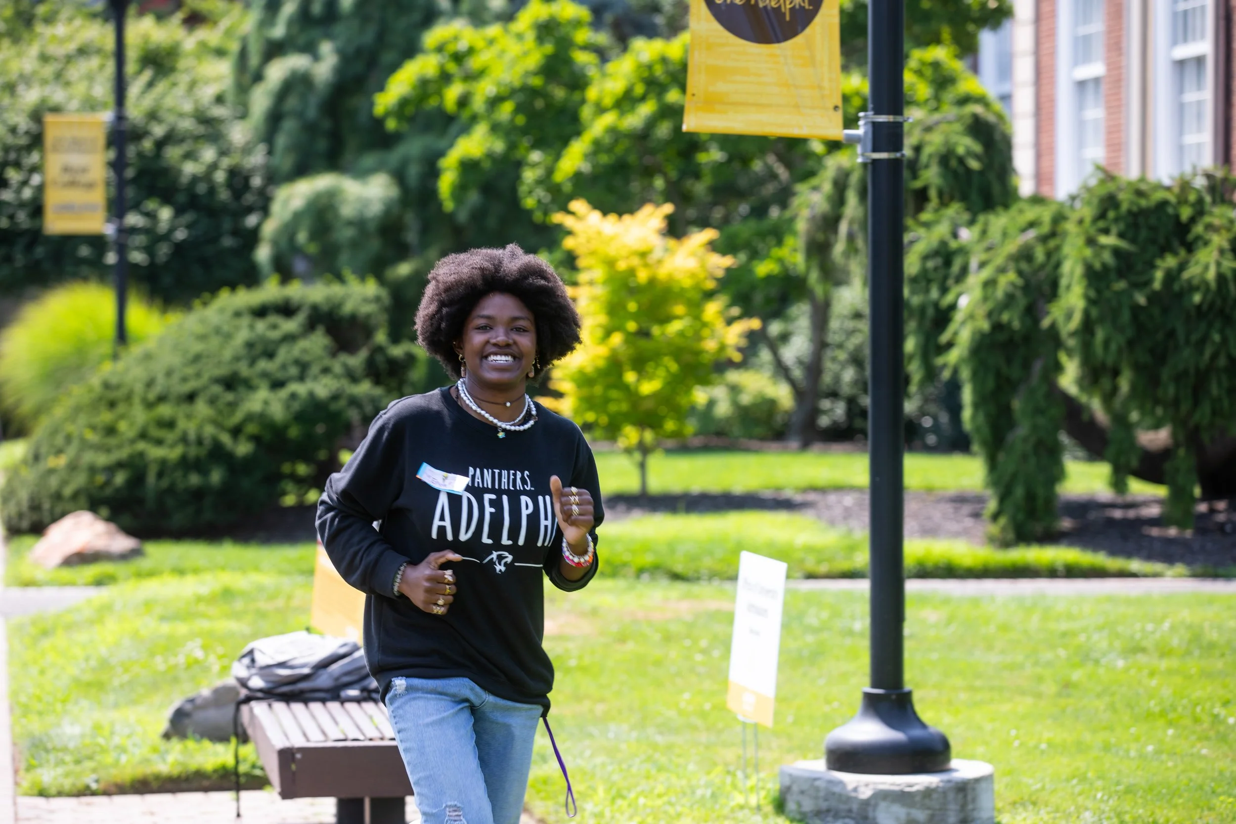 A smiling woman wearing a black Adelphi University sweatshirt and jewelry, jogging outdoors in a green park on a sunny day.
