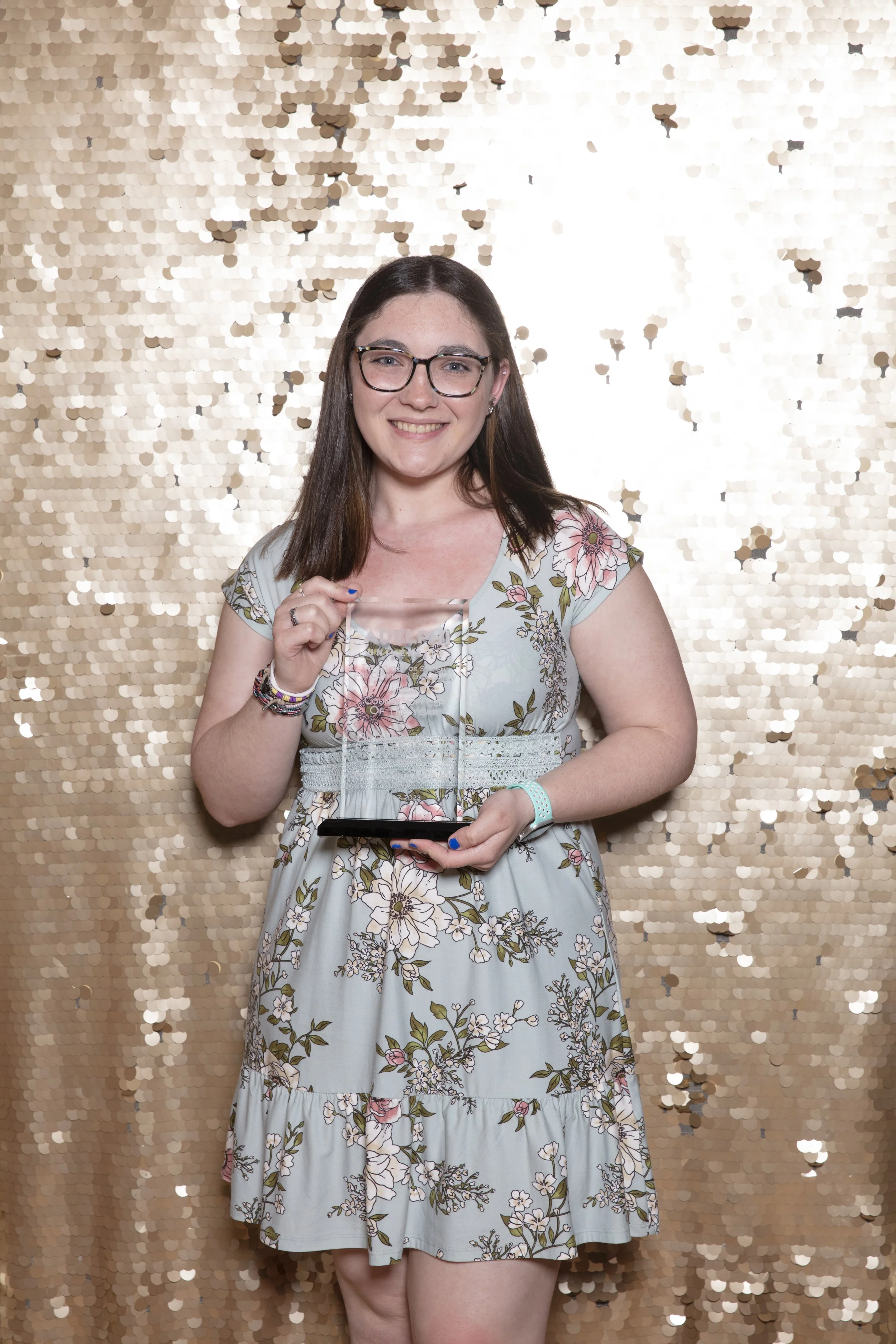 A young woman with glasses and long brown hair smiling, holding an award and a black plaque in front of a gold sequin backdrop.