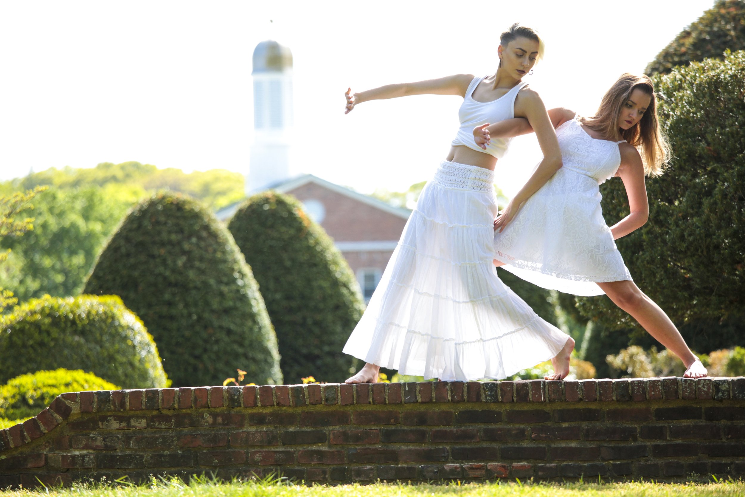 Two women in white dresses practicing yoga on a brick platform outdoors, with green bushes and a building with a clock tower in the background.