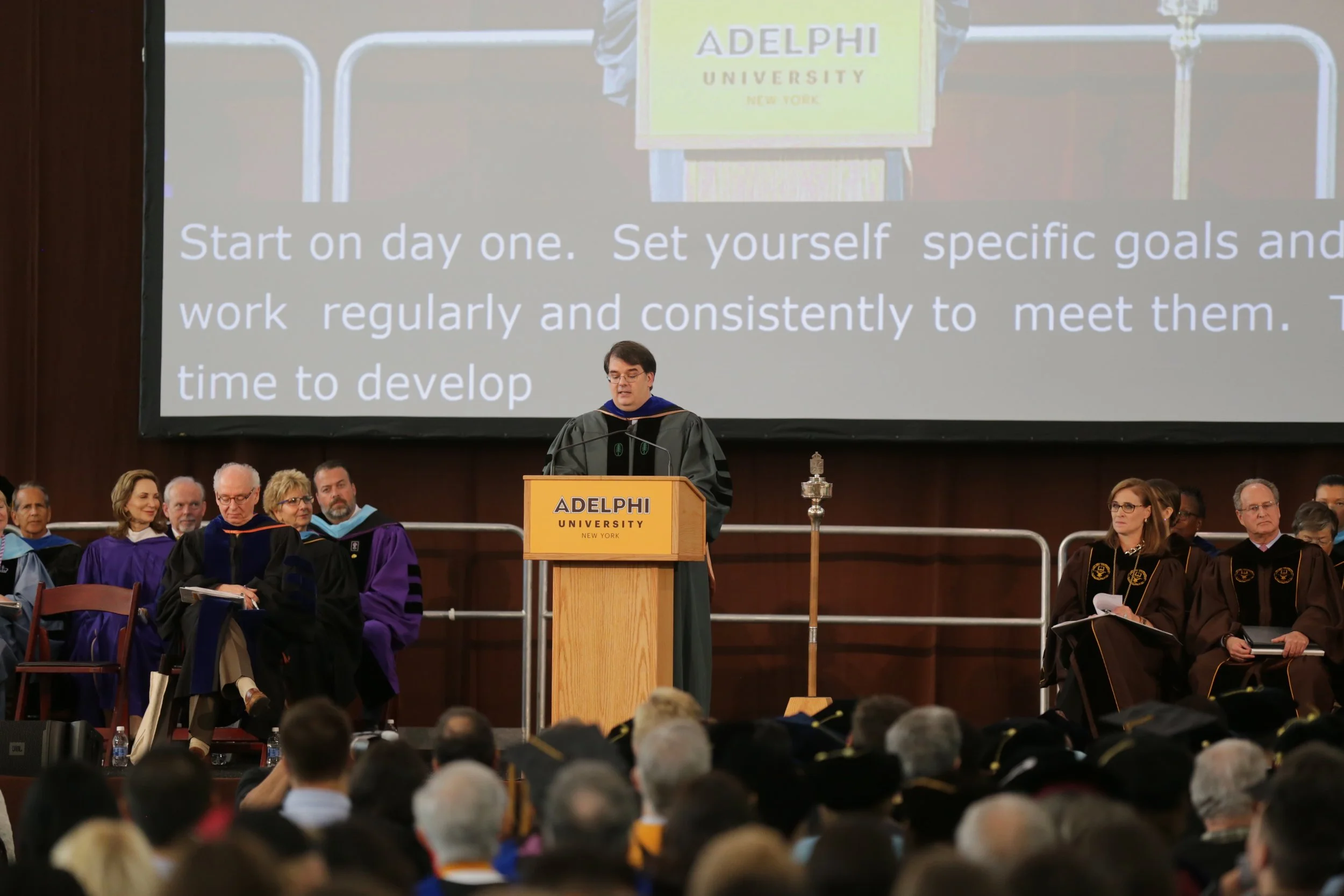 Graduation ceremony at Adelphi University in New York, with a speaker at the podium and faculty seated behind. The audience is visible in the foreground.