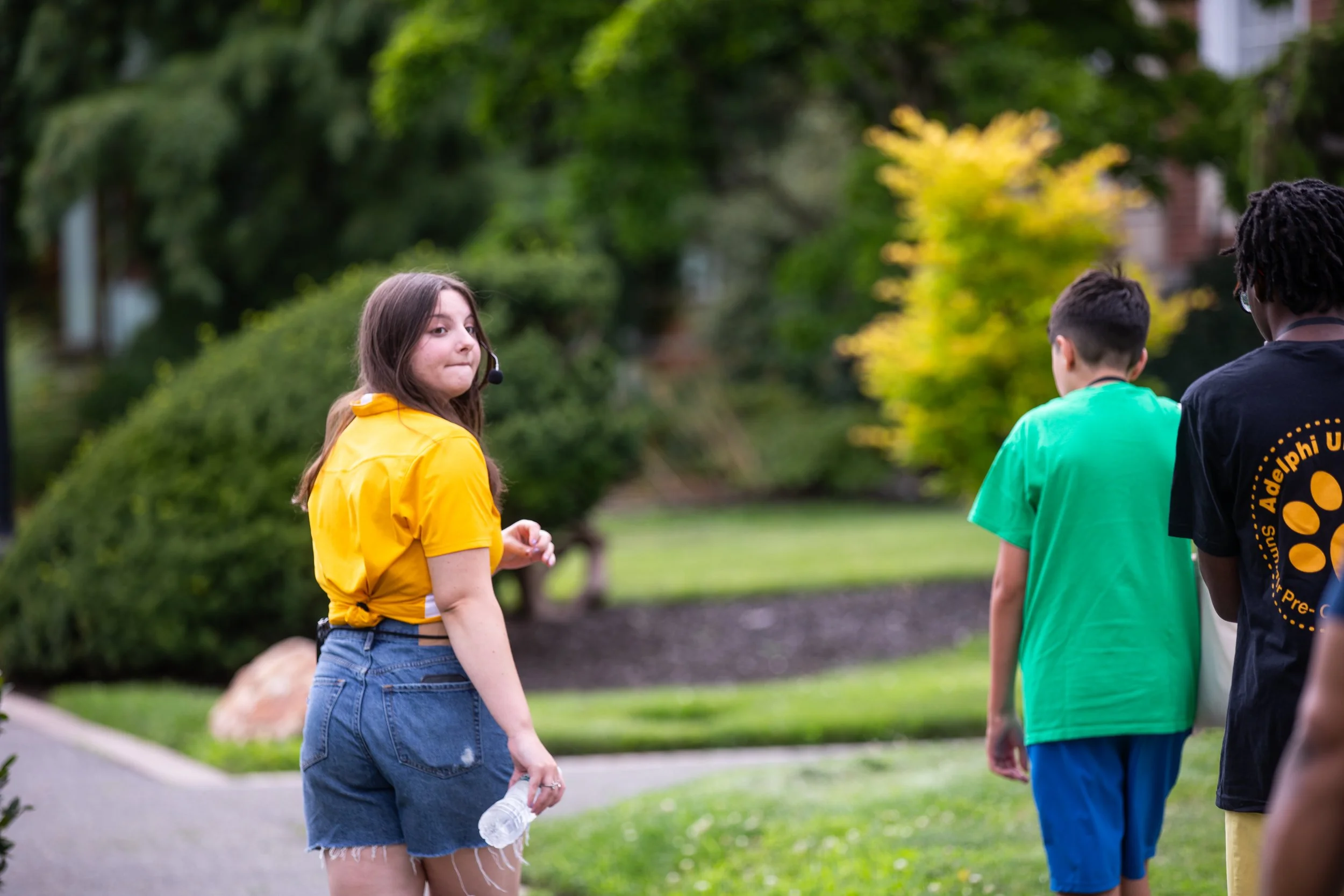 A young woman with long brown hair, wearing a yellow shirt and denim shorts, is standing outdoors in a park, holding a water bottle, with two boys in green and black shirts walking away from her in the background.