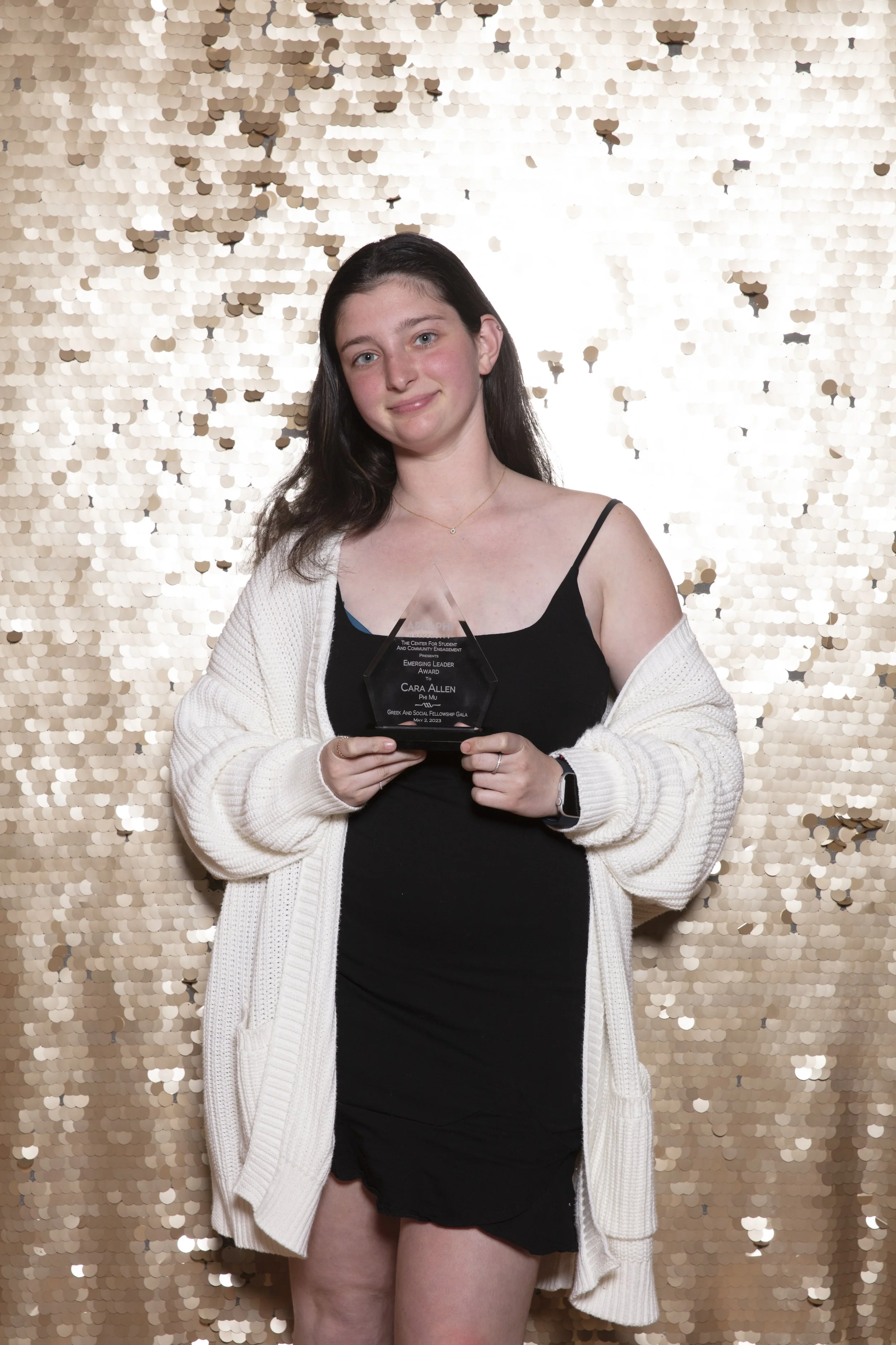 A young woman with long dark hair holding an award in front of a gold sequin backdrop. She is wearing a black dress, a white cardigan, and a smartwatch, smiling at the camera.