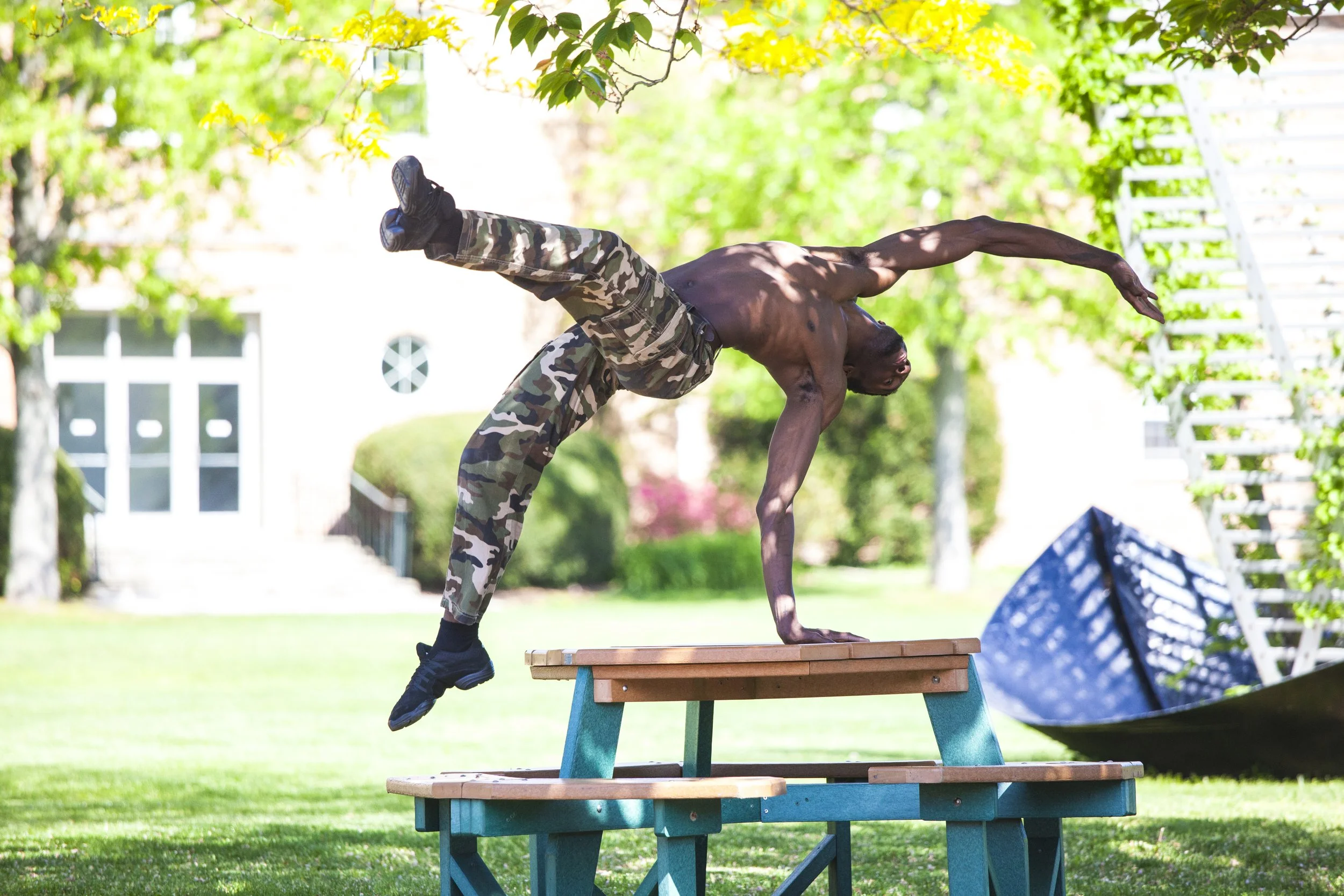 A shirtless man wearing camouflage pants and black shoes performing a handstand on a wooden table outdoors in a green yard with trees and a white building in the background.