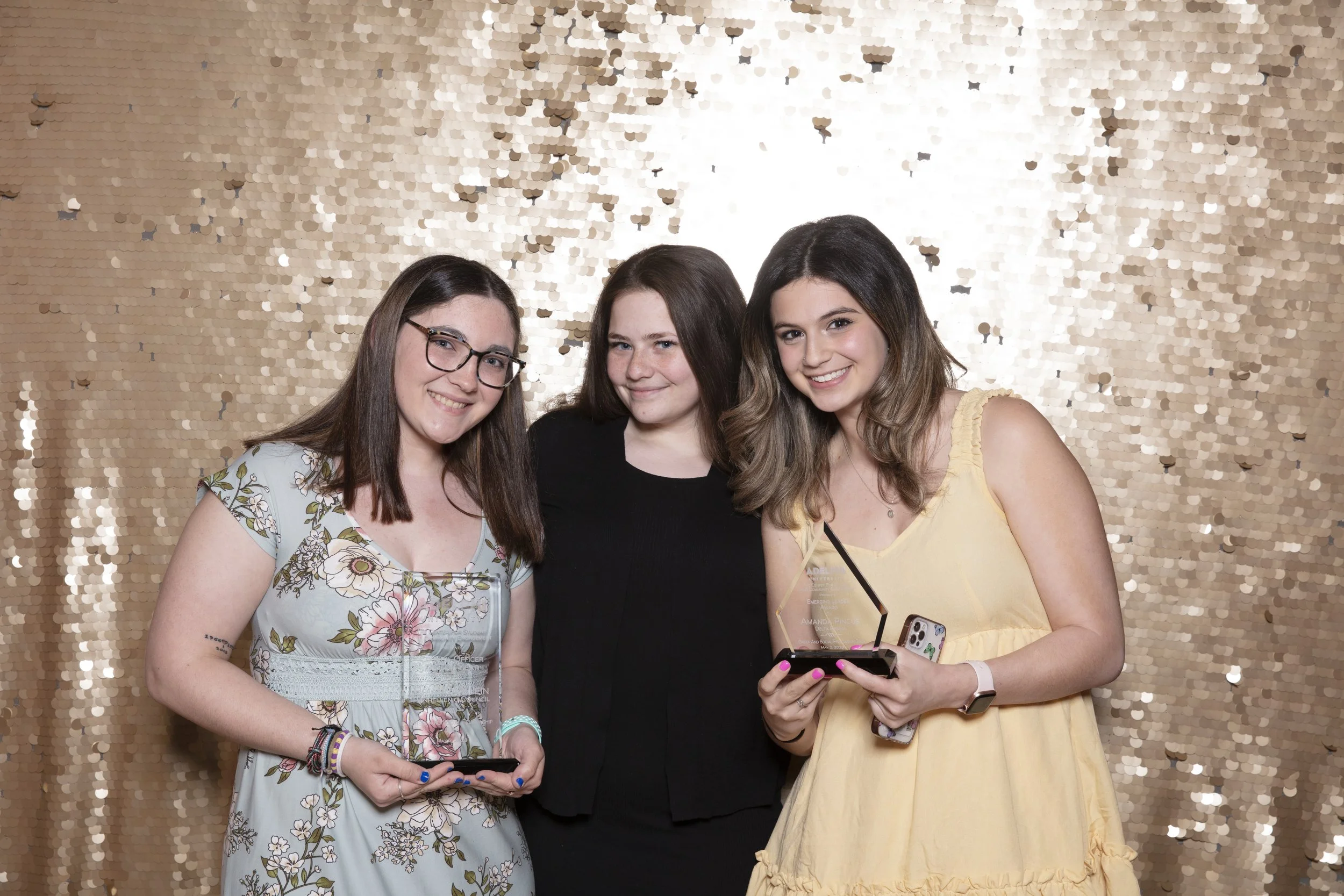Three young women smiling and holding awards and phones in front of a gold sequin backdrop at a celebration.