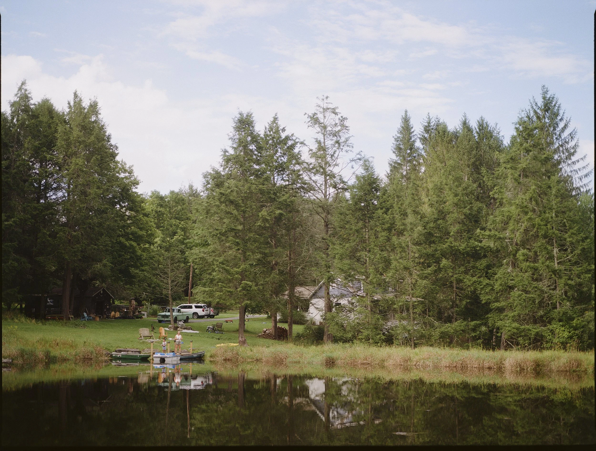 A peaceful lakeside scene with children playing near the water, boats docked at the shore, and trees in the background across a calm lake.