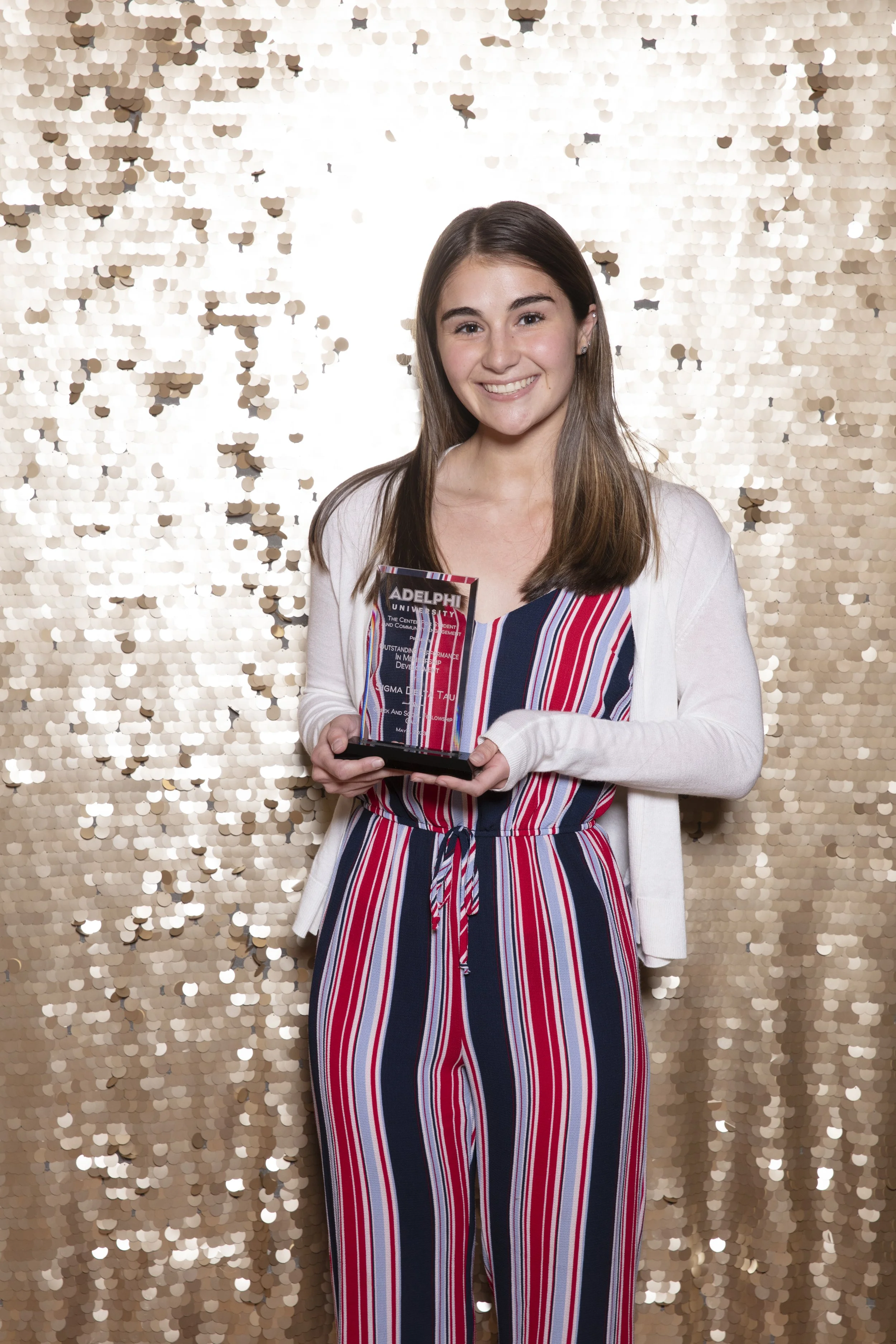 Young woman holding an award trophy in front of a gold sequin backdrop, smiling, wearing a striped jumpsuit with a white cardigan.