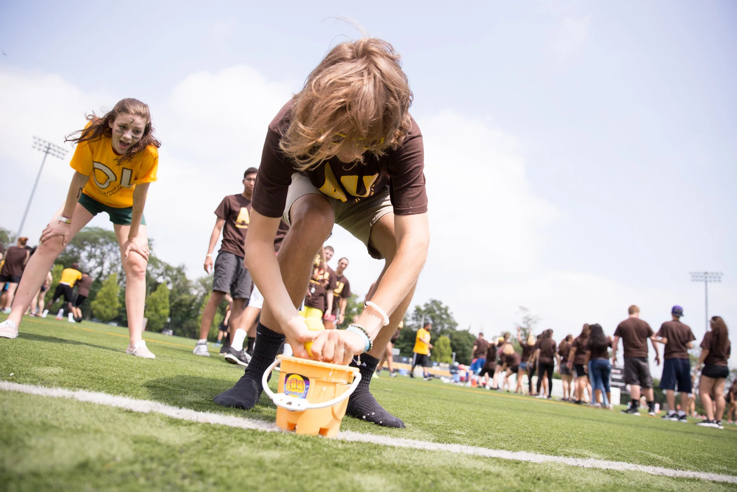 Teenager with long hair crouching on a sports field, digging a yellow bucket in the grass while others watch, some in school jerseys, outside on a cloudy day.