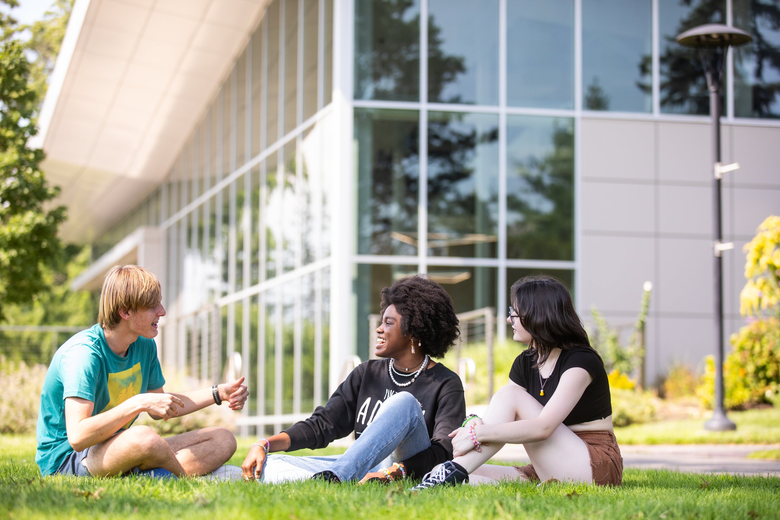 three young people sitting on grass outside, conversing and smiling, near a modern building with large glass windows, surrounded by trees and a lamppost.