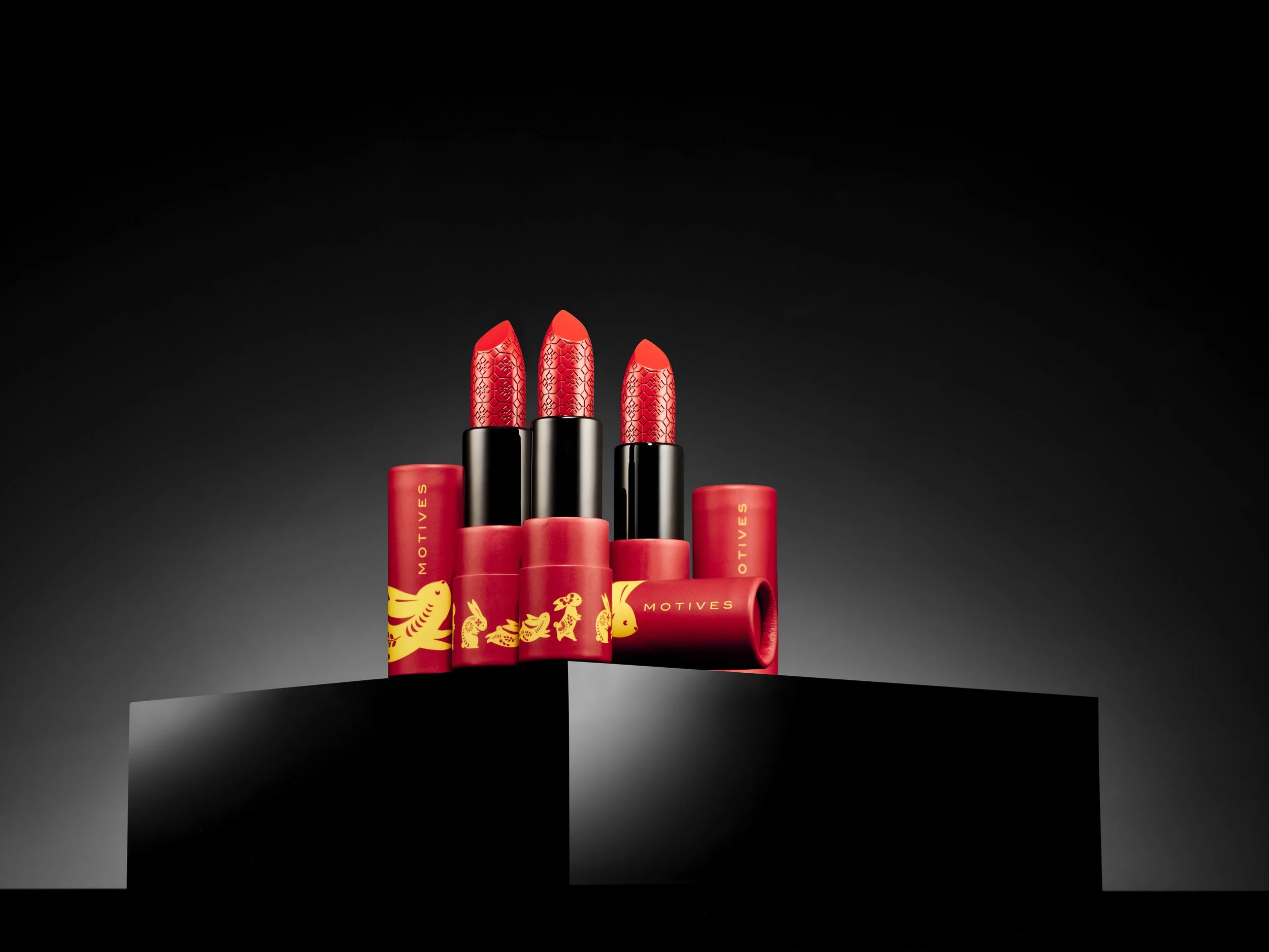 Three red lipsticks with gold dragon and rabbit designs on the cases, displayed on a black pedestal against a dark background.