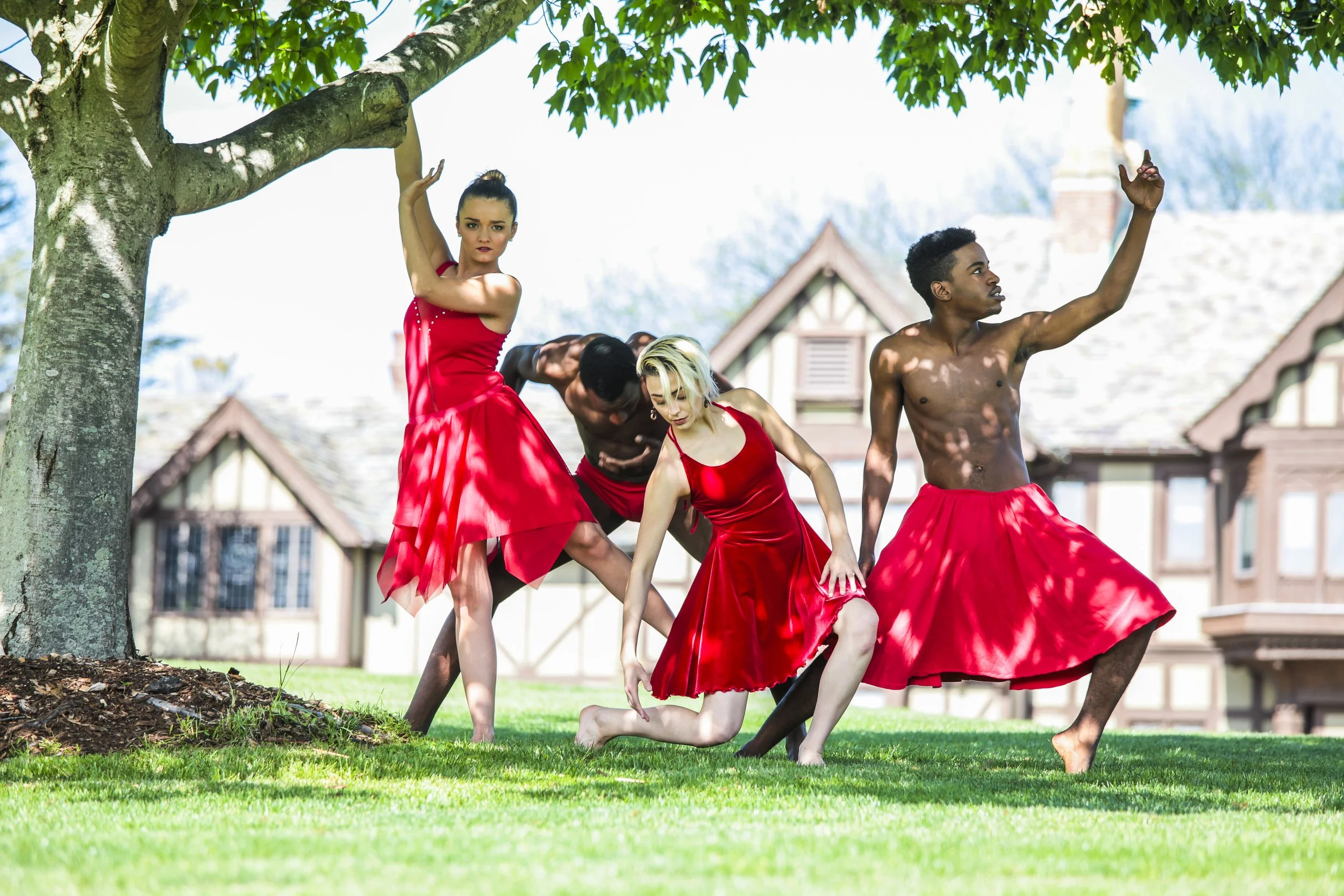 Four dancers are performing outdoors under a tree, wearing red dresses or skirts, with two men shirtless and two women in red dresses with different styles. They are posed dynamically on a grassy lawn with houses in the background.