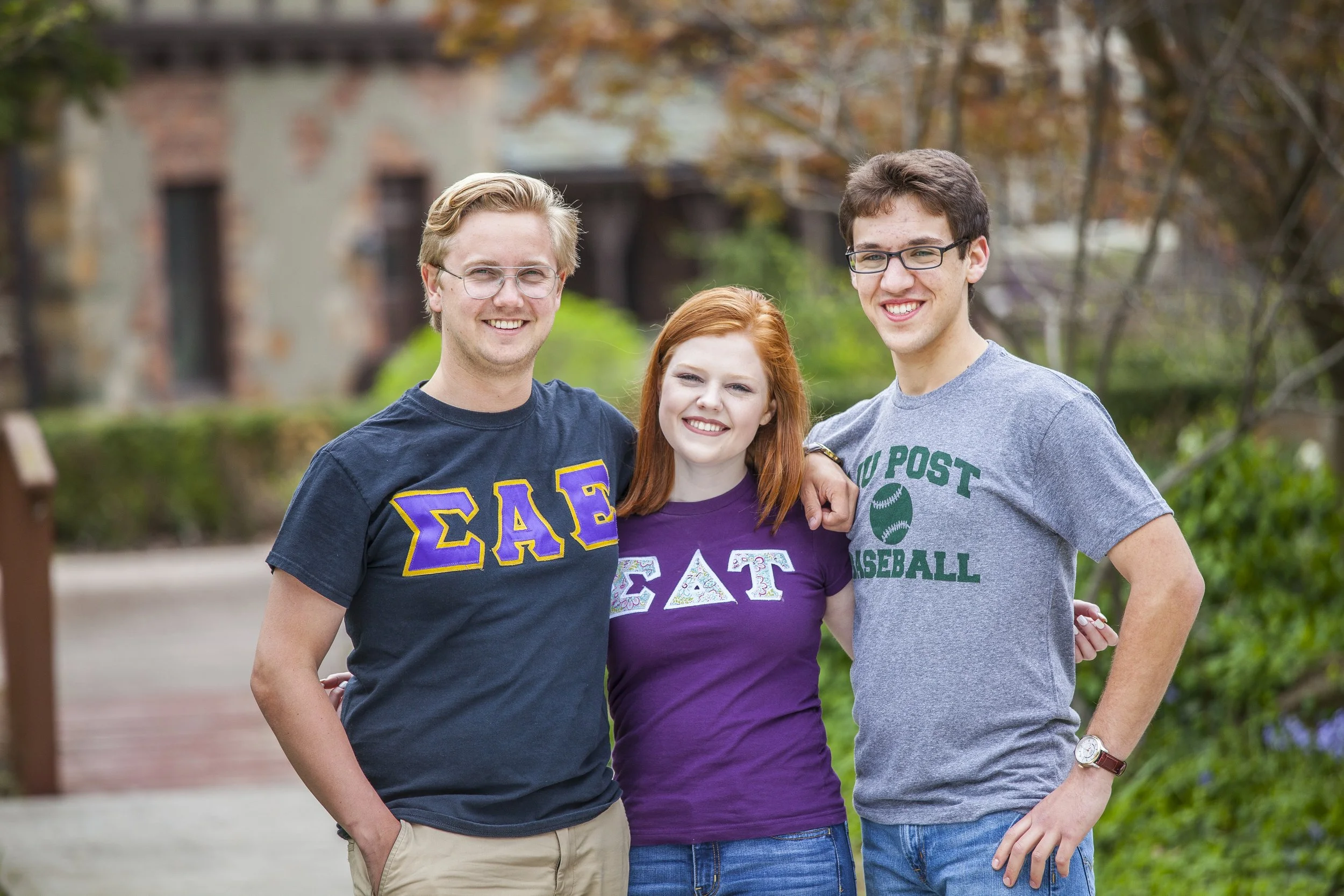 Three college students smiling outdoors with trees and buildings in the background, wearing Greek life and sports T-shirts.