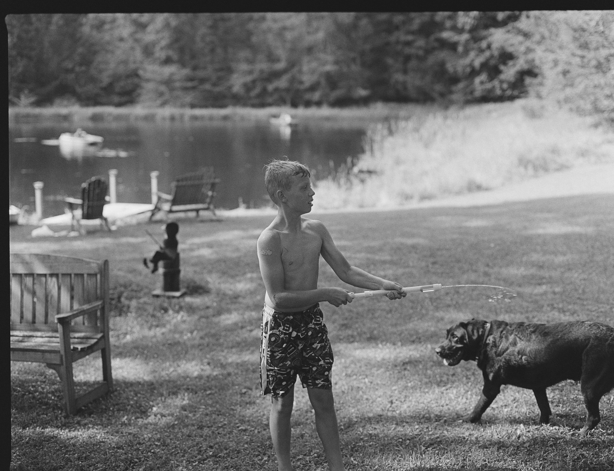A young boy with a tattoo on his arm and wearing patterned swim trunks playing with a dog by a lake, with boats and trees in the background.